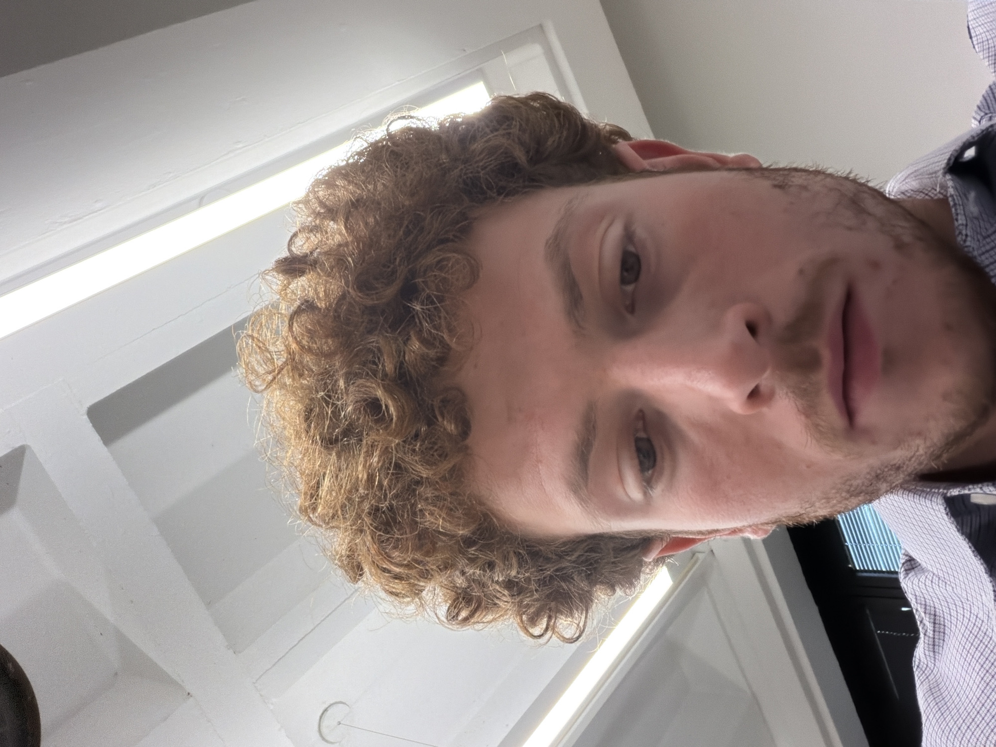 A low-angle, close-up photograph depicts a man with curly brown hair and a light complexion. He is wearing a button-down shirt with a subtle checkered pattern. The background includes a white ceiling with linear light fixtures, suggesting an indoor environment, possibly an office. The location is impossible to determine based on the image alone. The man is looking directly at the camera.