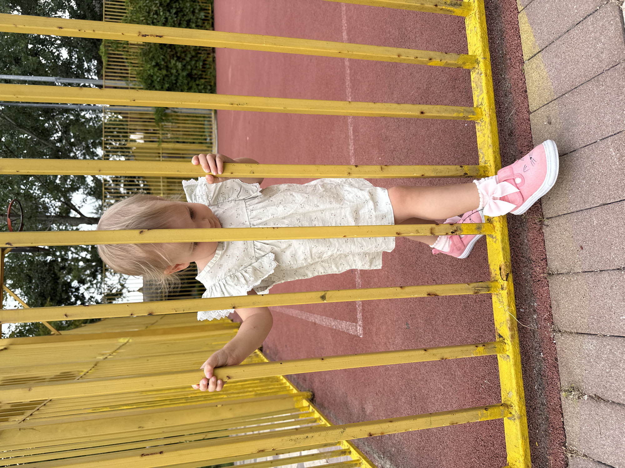 A young Caucasian female child with blonde hair, wearing a white dress and pink shoes, stands behind a yellow metal fence. The child is looking down and holding onto the fence. The background shows a red sports court with white markings, trees, and a green hedge. The scene appears to be an outdoor playground or park.