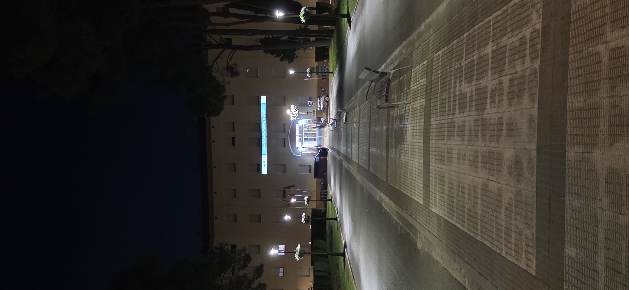A night time, eye-level, vertical, full shot shows the exterior entrance of a hospital at the end of a pathway. The building is light beige with multiple rows of square windows. Above the entrance, a lit blue sign reads "SALUD SAGRADO CORAZÓN DE JESÚS". The entrance itself is an archway with glass doors. Flanking the path are lampposts with hanging flower baskets. Three benches are visible along the pathway. To the right, trees form a dark silhouette against the night sky. The pathway is paved with light-colored rectangular tiles with dark grout, and the sides of the path are lined with dark gray concrete. A red van is parked to the left of the entrance, and a silver car is parked to the right. The scene is in Huesca, Spain.