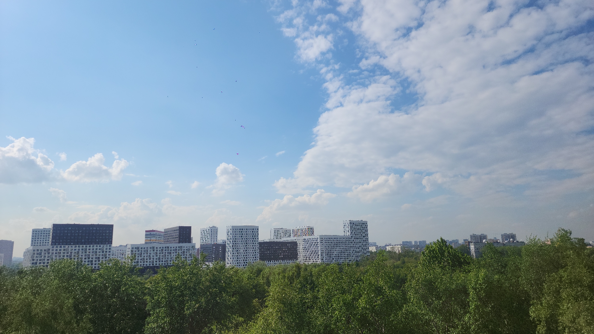 A sunny cityscape in Moscow, Russia, features a line of modern apartment buildings nestled behind a dense green canopy of trees. The buildings are predominantly white with varied dark accents, displaying a repetitive pattern of windows and balconies. In the sky, a gradient of blue is dotted with scattered white clouds and several small purple objects, possibly balloons, float high above. The lush green foliage in the foreground suggests a park-like setting.