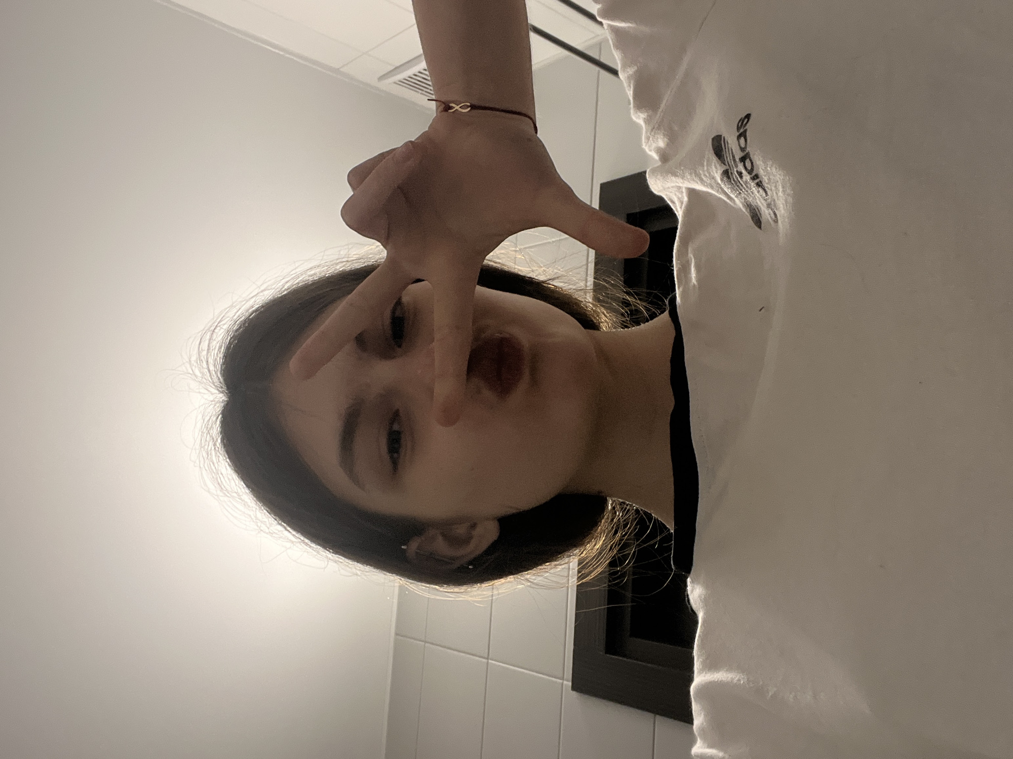 An indoor selfie features a young individual, likely female, with dark hair, playfully posing for the camera. The person is making a "V" sign hand gesture (peace sign) with their left hand, partially obscuring their face, while pouting their lips. They are wearing a white t-shirt with a black "adidas" logo visible on the upper right side. A red string bracelet adorned with a gold-colored infinity symbol is on their left wrist.
The setting appears to be a room with white rectangular wall tiles in the background, possibly a bathroom or utility area. A dark rectangular frame, which could be part of a mirror or a window, is visible behind the person's right shoulder. A ceiling vent is present in the upper left of the background. The image is taken from a low angle, looking upwards.
A prominent feature is the strong backlighting, creating a bright halo effect around the subject's head and causing parts of their face to be in shadow. The overall emotion conveyed is playful and casual. The image is originally rotated 90 degrees clockwise. The location context is Unknown, Russia.