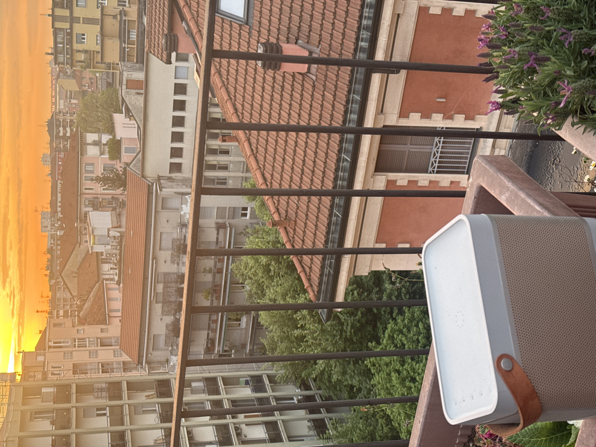 The scene depicts a view from a balcony in Milan, Italy. The image includes buildings with terracotta roofs, balconies, and green trees, characteristic of the urban landscape. A black metal railing runs horizontally across the frame, partially obscuring the view. In the foreground, a white speaker sits on the balcony, positioned near a flower pot with purple flowers. The sky is visible in the distance, displaying a golden hue, suggestive of a sunset. The buildings are of various heights and architectural styles.