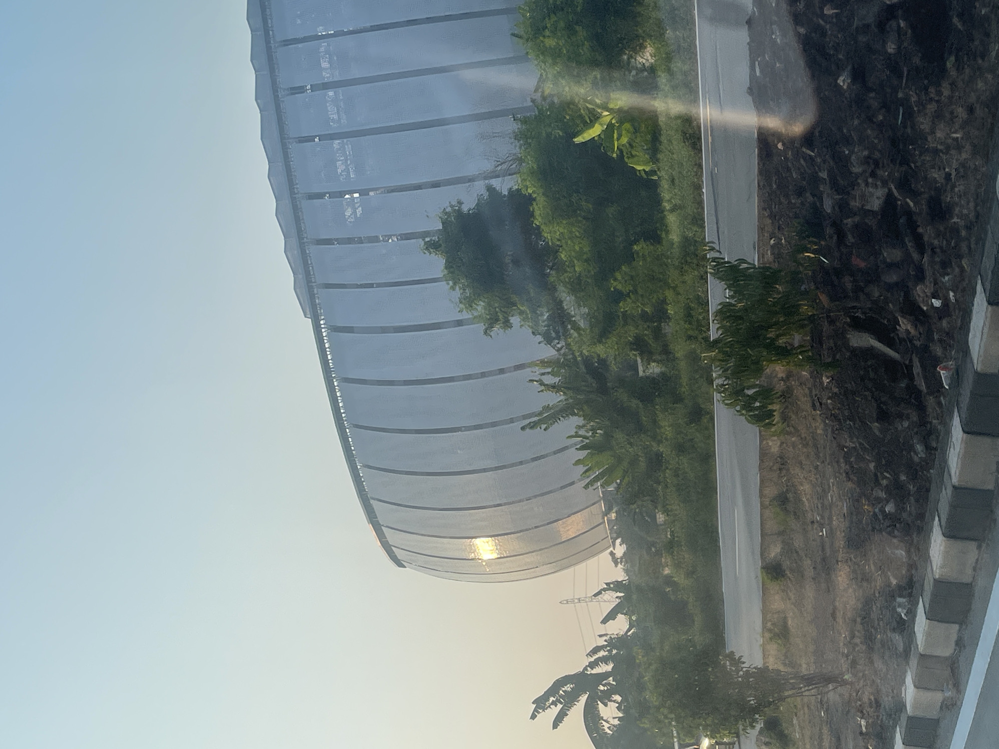 The image shows the exterior of the Jakarta International Stadium, a large, modern stadium with a distinctive curved roof covered in white panels. The sun is shining brightly, reflecting off some of the panels, creating a warm glow. The stadium is surrounded by lush green trees and vegetation, suggesting a tropical environment. In the foreground, there is a road and a strip of land with some debris and small plants. The sky is clear and pale blue. The overall impression is of a grand, contemporary structure set against a natural backdrop on a sunny day.