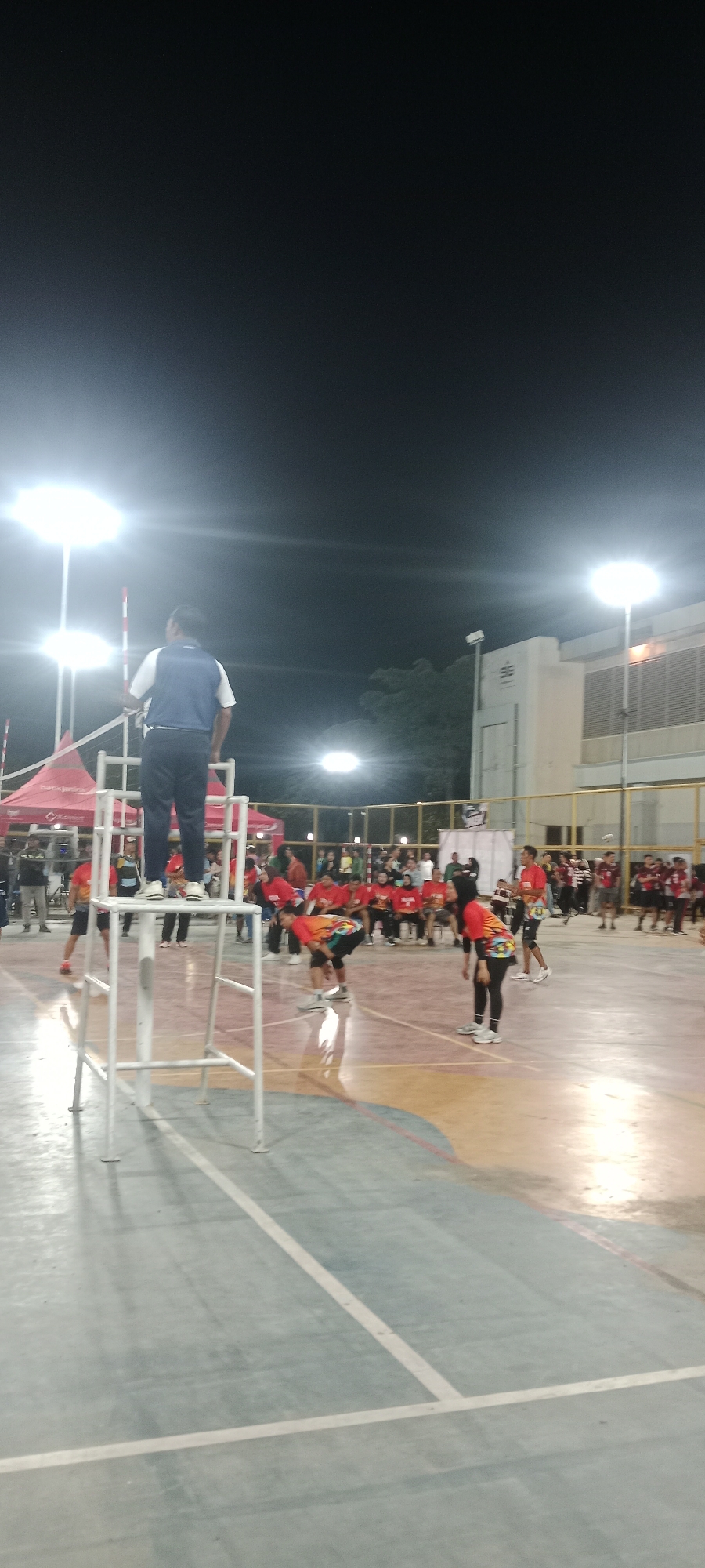 The image depicts a night scene at an outdoor volleyball court, likely in the city of Tuban, Indonesia, given the context provided. A man in a blue and white shirt and dark pants stands on a raised platform, observing a volleyball game. Several individuals, mostly wearing red shirts, are positioned on the court, some in a defensive stance, suggesting an active match. Bright floodlights illuminate the court from above, casting reflections on the polished floor. Tents with logos are visible in the background, along with a building structure. The atmosphere appears to be that of a local sporting event or competition.