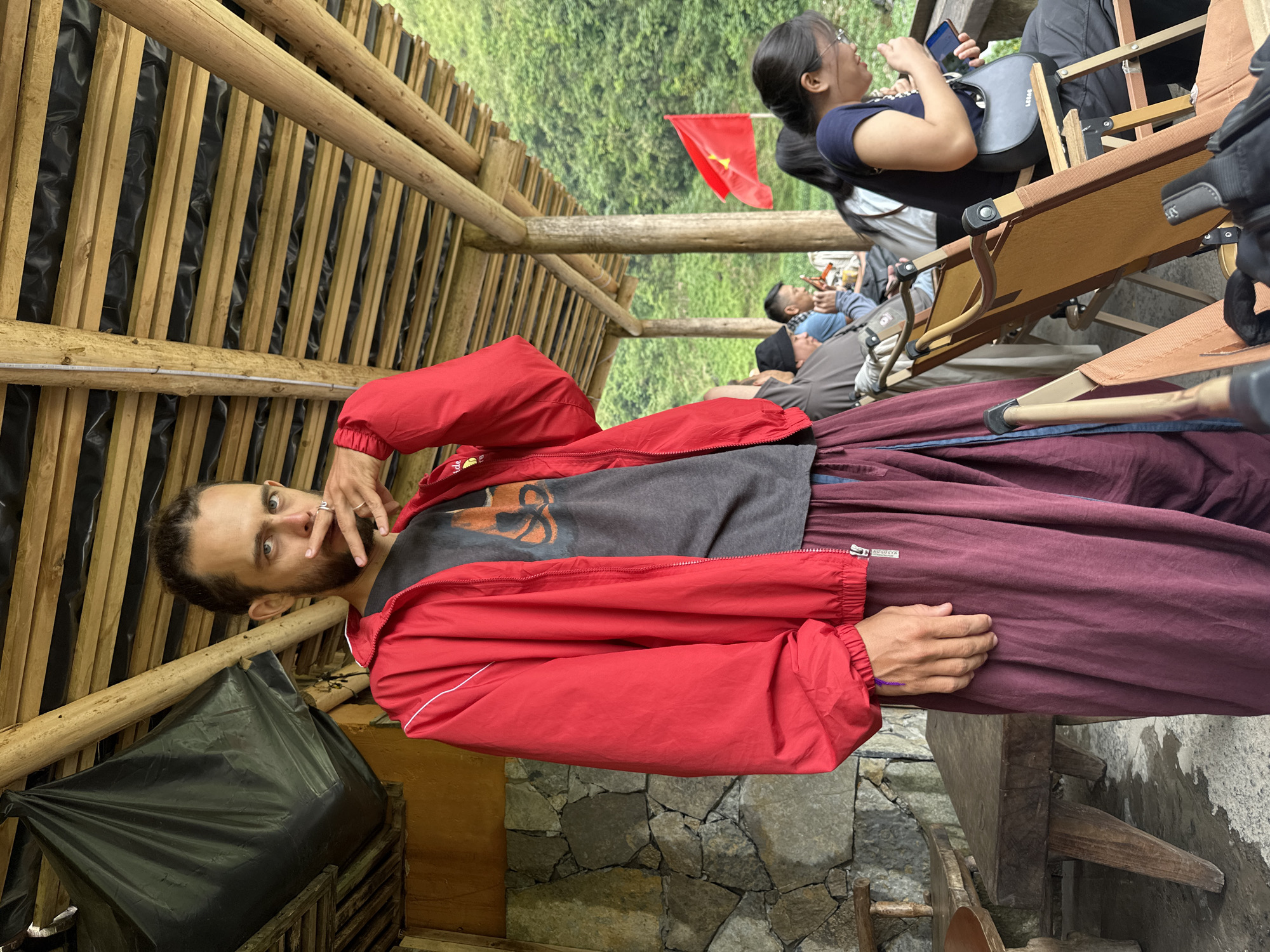 Captured in Yên Minh Commune, Vietnam, the photograph presents a man in a red jacket and maroon pants, positioned in a structure with bamboo-like supports and wooden crossbeams. The setting suggests an outdoor or semi-enclosed space, possibly a shelter or seating area. He is seen in the center. There is a Vietnamese flag in the background, along with other people sitting and using mobile devices. A black trash bag is nearby and the structure seems to be built from wood. The wall is made of stacked stones.