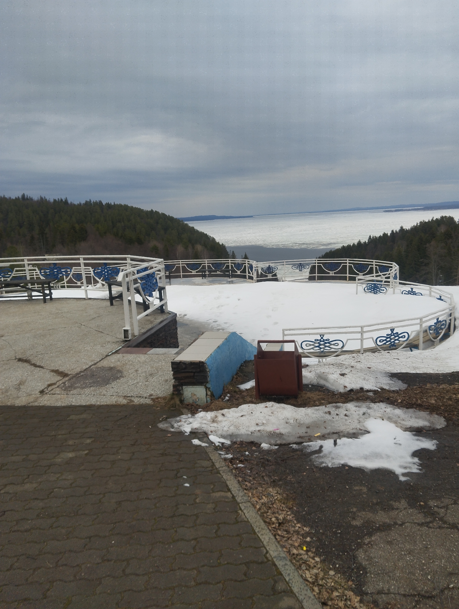 The image shows an outdoor viewing area in Galevo, Russia, likely in early spring due to the presence of melting snow. The area features a raised concrete platform with benches and a decorative white metal railing with blue accents. In the background, a body of water covered with ice floes can be seen, bordered by forested hills. The sky is overcast. In the foreground, there are patches of snow and debris, a blue concrete structure, and a brown trash receptacle. The ground is paved with interlocking pavers. There are no people visible in the image.