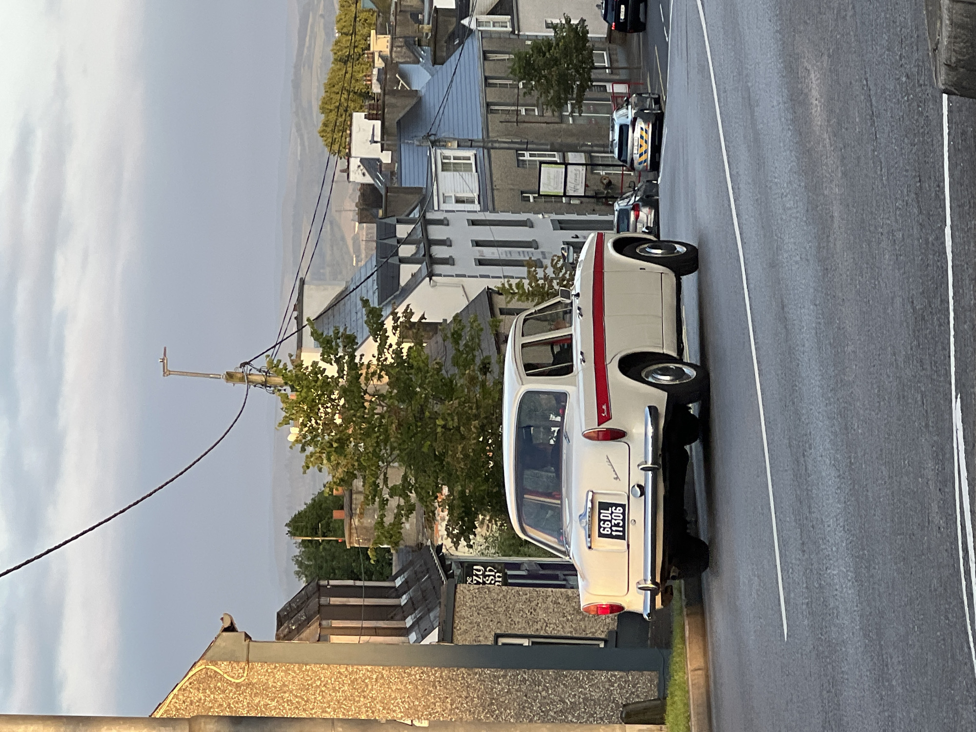 A cream-colored vintage sedan with red racing stripes on the sides is parked on a street in Mountcharles, Ireland. The car has the license plate "66 DL 11306". In the background, several buildings line the street, some with blue roofs and others with grey stone facades. Trees are visible, and the sky is overcast with hints of distant hills. Two other vehicles, a dark-colored SUV and a silver car, are also present on the street, with the silver car appearing to be a police vehicle based on its markings. The street is paved, and a white line marks the edge of the road. The scene appears to be captured during daylight, possibly late afternoon given the lighting conditions.