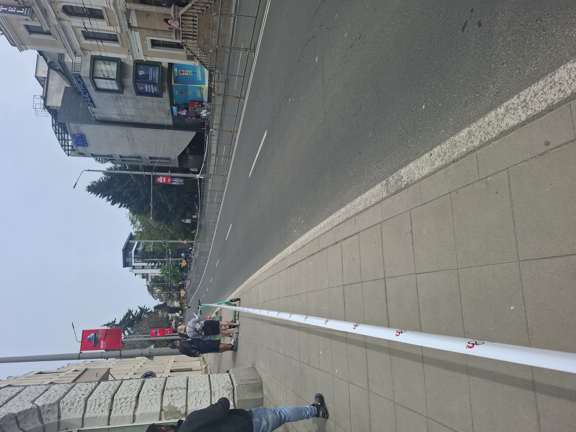 The image captures a street scene in Burgas, Bulgaria. The view is along a wide street with a well-maintained asphalt surface and a white dividing line. A gray sidewalk runs along the right side of the street. A white barrier tape with red logos stretches across the sidewalk. Two people stand near a green electric scooter, seemingly interacting with it. Another person is partly visible in the foreground, wearing jeans and dark shoes. Buildings line the left side of the street, some with visible signage and architectural details. Red banners are also visible on poles along the street. The sky appears overcast.