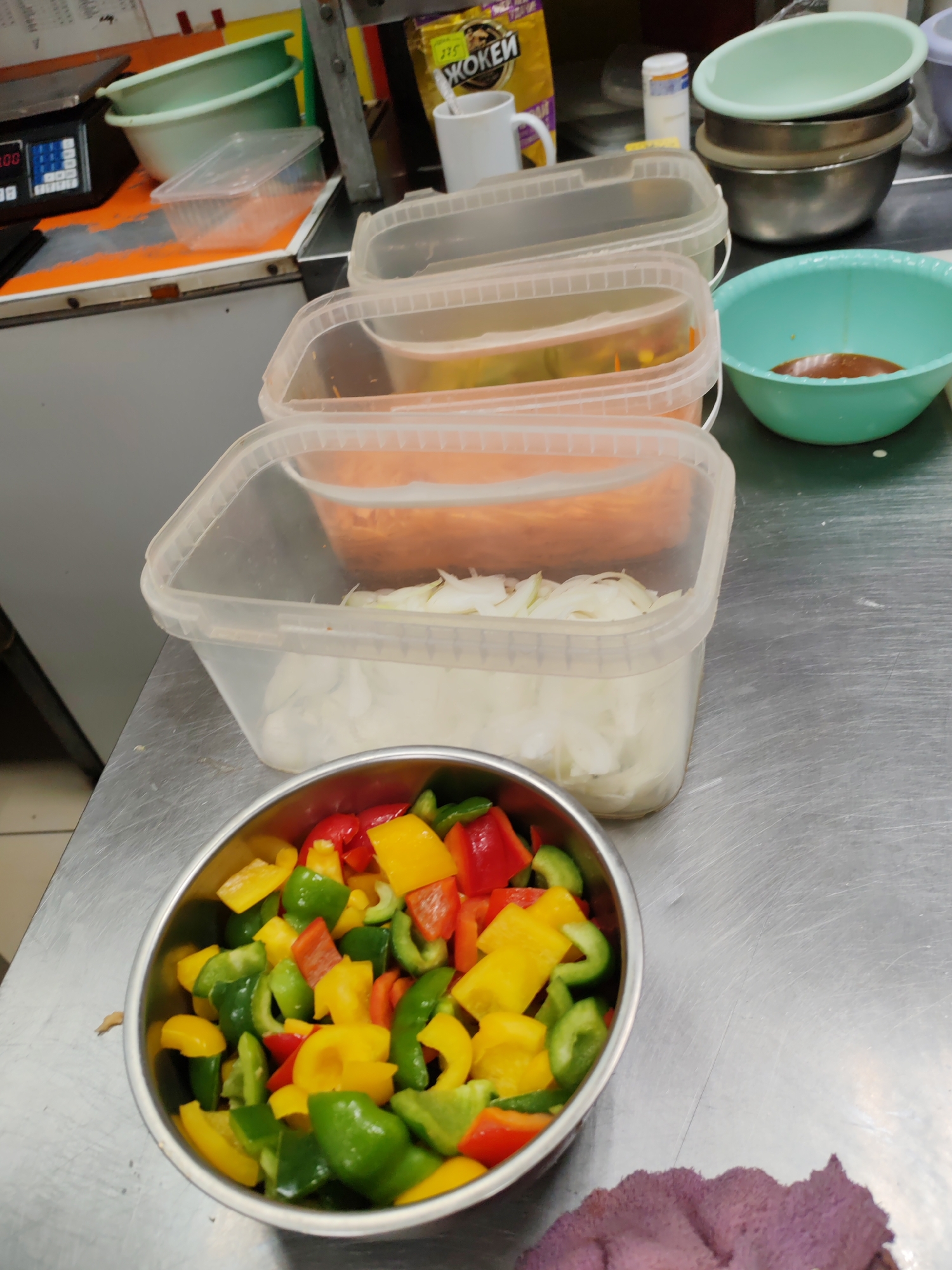 In Komsomolsk-on-Amur, Russia, a stainless steel countertop in what appears to be a commercial kitchen environment displays an assortment of food preparation items. Prominently featured is a metal bowl filled with chopped bell peppers in red, green, and yellow. To the right of the bowl are three clear plastic food storage containers, stacked and partially filled. The top container holds chopped white onions, the middle contains shredded carrots, and the bottom is obscured by the others but likely holds similar ingredients. Further to the right, a green plastic bowl contains a dark, viscous liquid, possibly a sauce or marinade. In the background, several stacked metal and plastic bowls are visible, along with a white mug and a yellow packet of "Жокей" (Jockey) coffee. A digital scale with a black base and a display showing "0.00" is on the left side of the frame, positioned on an orange and white surface. A gray wall forms the backdrop. The overall scene suggests an ongoing food preparation process.