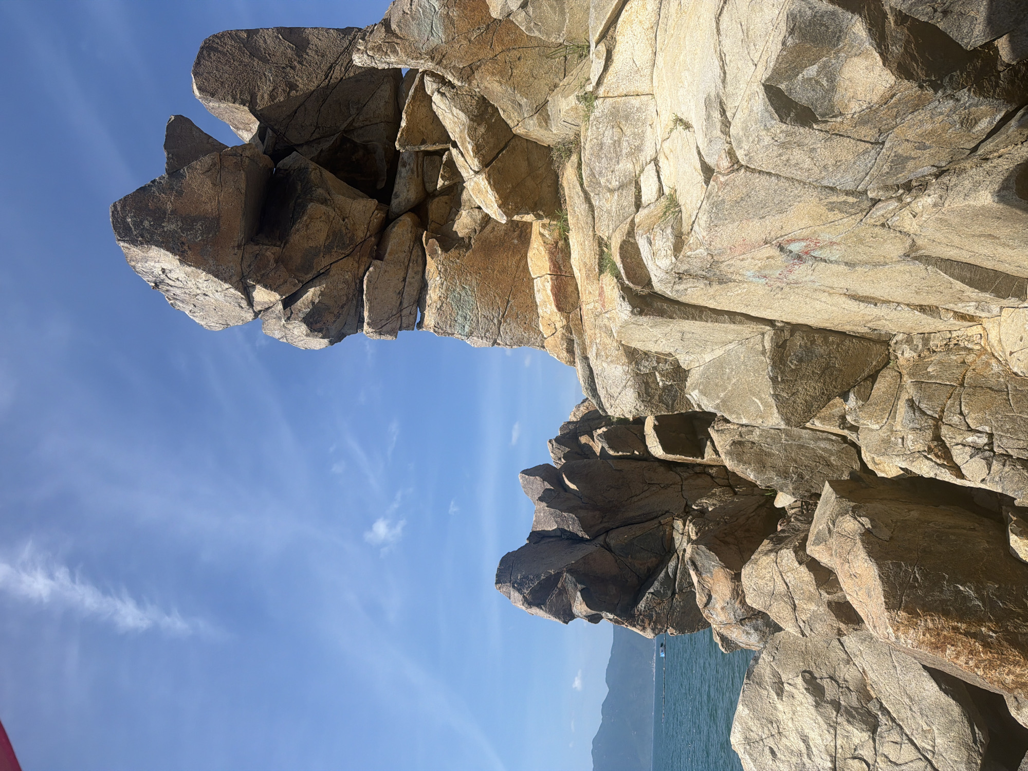 This image captures a section of the coastline in Vĩnh Lương, Vietnam, characterized by large, weathered rock formations against a clear blue sky with scattered white clouds. The rocks are a mix of tan, brown, and grey, with visible cracks and fissures indicating geological activity and erosion. Some rocks are stacked precariously, creating a rugged and dramatic landscape. Green vegetation sprouts from some of the crevices in the rocks. In the background, the turquoise ocean stretches towards a hazy mountain range. A small, dark-hulled boat is visible on the water to the left of the frame. The lighting suggests it is daytime, with sunlight casting shadows and highlighting the textures of the rock surfaces.