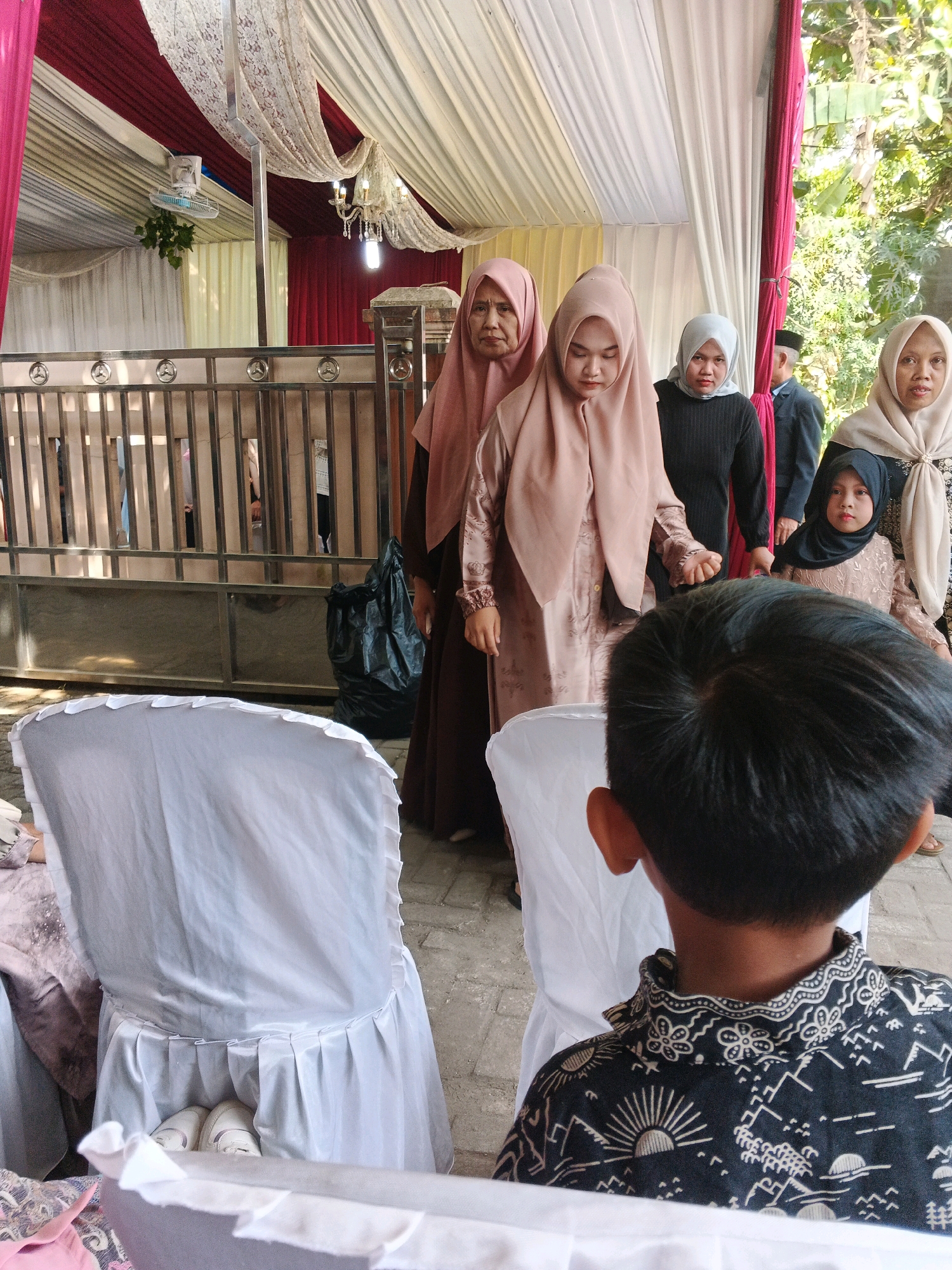 In a covered outdoor area, possibly a wedding venue, in Kadu, Indonesia, several women and a young girl, all wearing hijabs, stand in a line. The woman on the far left wears a pink hijab and a dark brown dress. The woman next to her, in the center, is dressed in a blush-colored hijab and dress. To her right, a woman in a light grey hijab and dark top is visible. On the far right, a woman in a beige hijab and a dress with intricate patterns looks towards the camera. A young girl in a black hijab and a light-colored dress stands in front of her. The backdrop includes a metal fence, draped fabric in white, maroon, and cream, and a ceiling fan. In the foreground, two white-covered chairs are prominent. A black trash bag is also visible near the fence. The overall scene suggests a formal gathering or event.