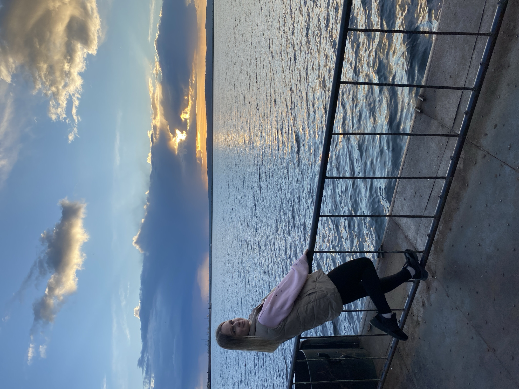 A young woman with long blonde hair sits on a black metal railing. Her body is rotated about 60 degrees clockwise. She is wearing black pants and a jacket, and she looks directly at the camera with a smile. Behind her is a large body of water that reflects the setting sun. The sky is blue with scattered clouds. Based on the information available, the location is Chaikovsky, Russia.