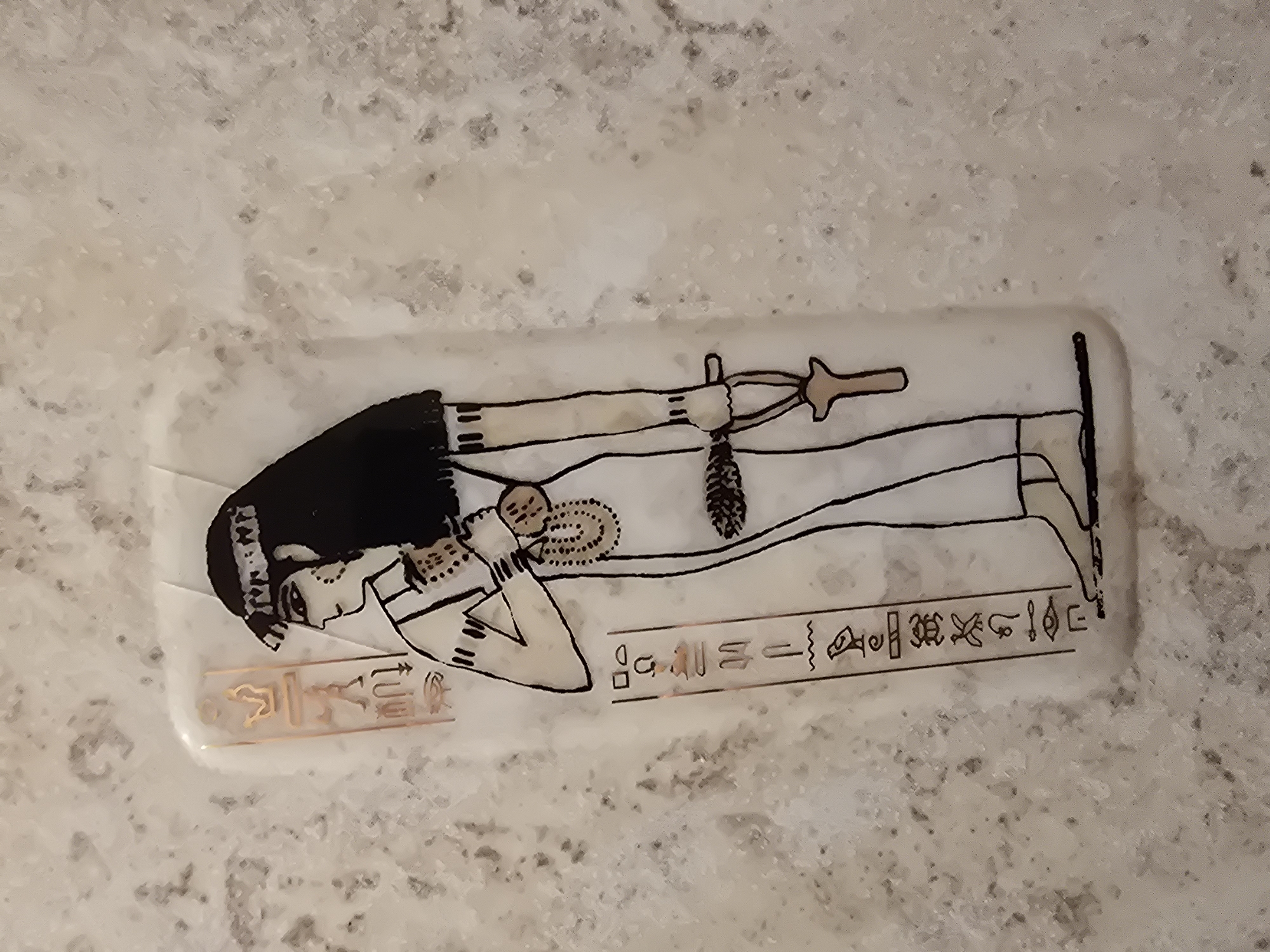 An Egyptian-themed decoration is displayed on a speckled, light-colored surface. The decoration features a depiction of a standing Egyptian figure in profile, facing left. The figure wears a headdress, a white dress, and a beaded necklace. They are holding an ankh symbol in their right hand and what appears to be a feathered object in their left. Hieroglyphs are present on both the left and right sides of the figure. The surface beneath the decoration has a textured, mottled appearance.