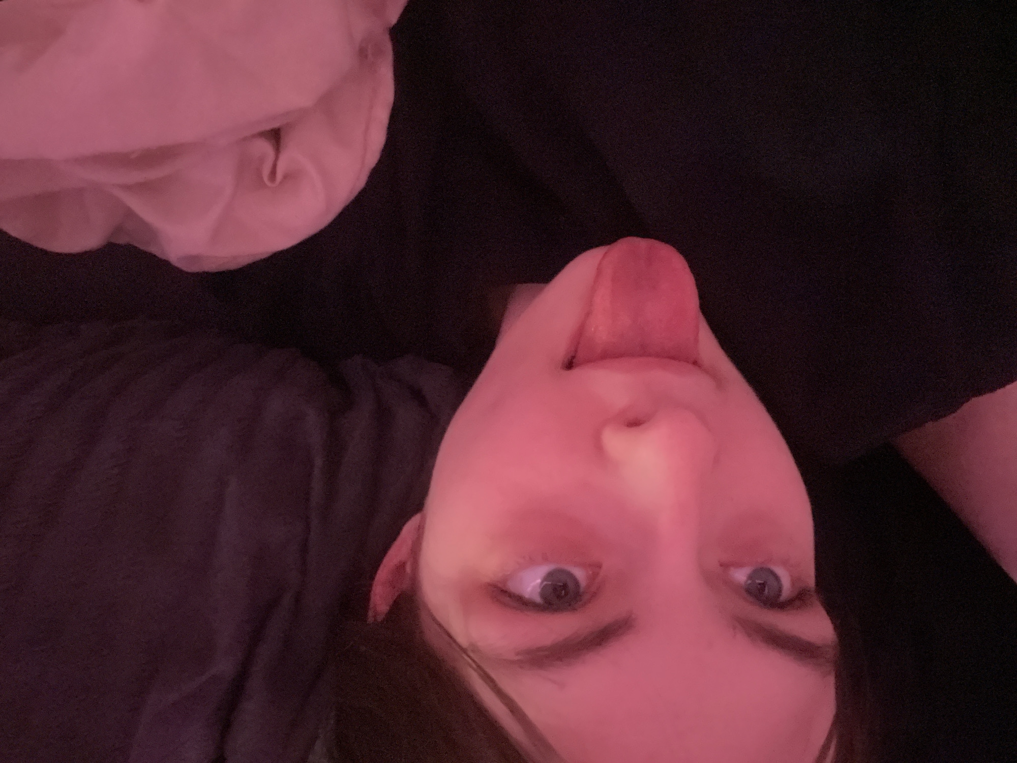A selfie taken from a low angle shows a person with their head tilted. The person has fair skin, dark hair, and blue eyes. Their tongue is sticking out. The background consists of dark fabric, possibly clothing or bedding, with a light-colored pillow or blanket in the upper left corner. The lighting appears to be predominantly pink. There is no information in the photo to suggest this picture was taken in Saint Petersburg, Russia.