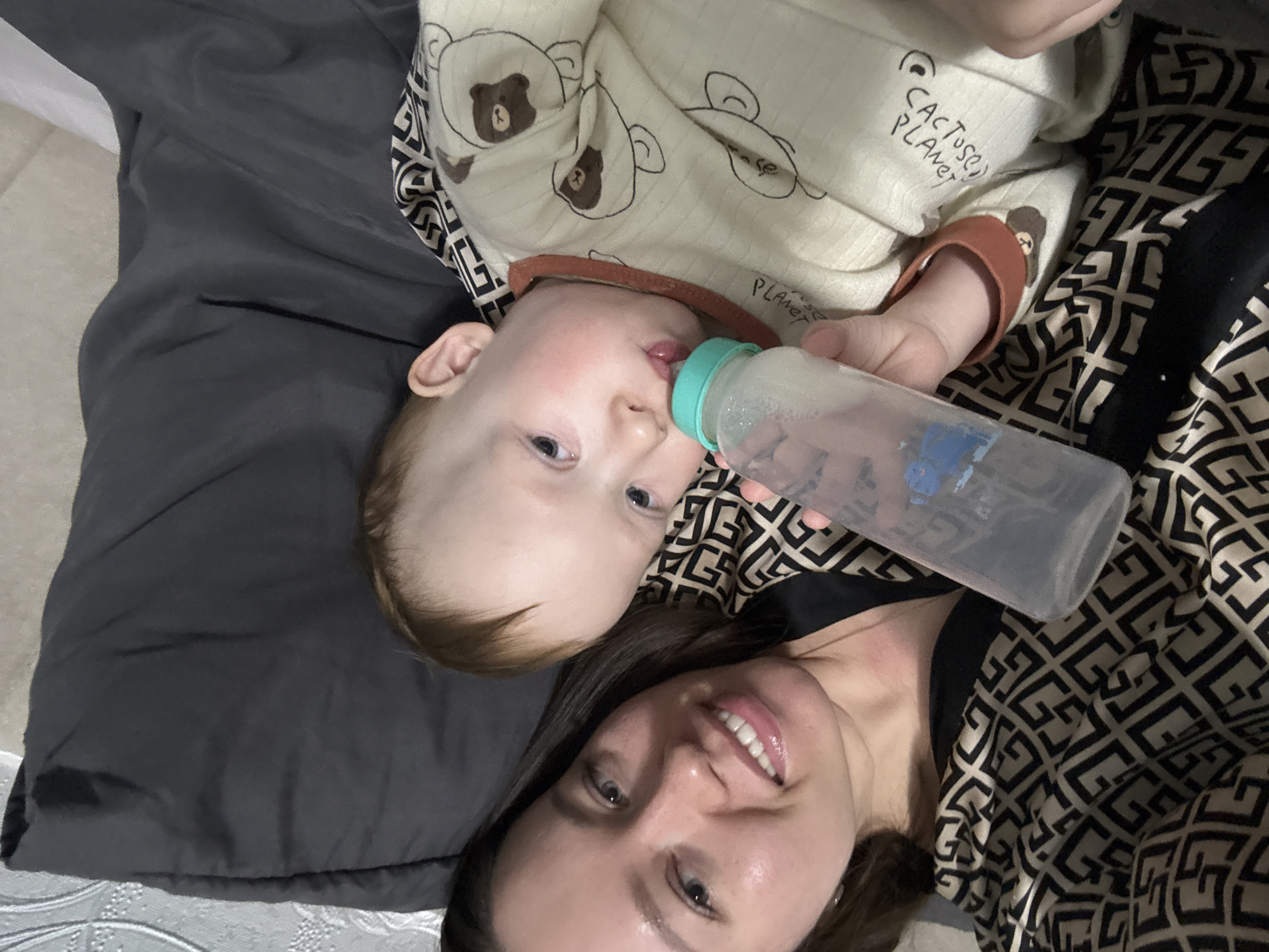 A woman and a baby are lying on a bed. The baby is positioned above the woman and is drinking from a bottle. The baby is wearing a light-colored patterned onesie, and the woman is wearing a black and patterned top. The bed has gray sheets. The photo was taken in Ферма, Russia.