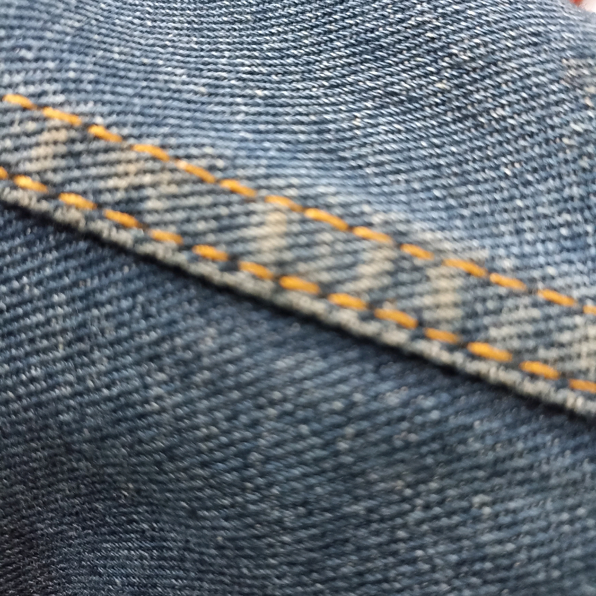 The image is a close-up, abstract shot of blue denim fabric with prominent orange stitching. The texture of the denim weave is visible, as are some lighter, faded areas on the fabric. The stitching runs diagonally across the image, creating a pattern of dashed lines. The lighting is even, and the focus is sharp on the texture of the fabric and the stitching. There are no people or identifiable objects in the scene, nor any indication of a specific location, including the city of Magelang, Indonesia.