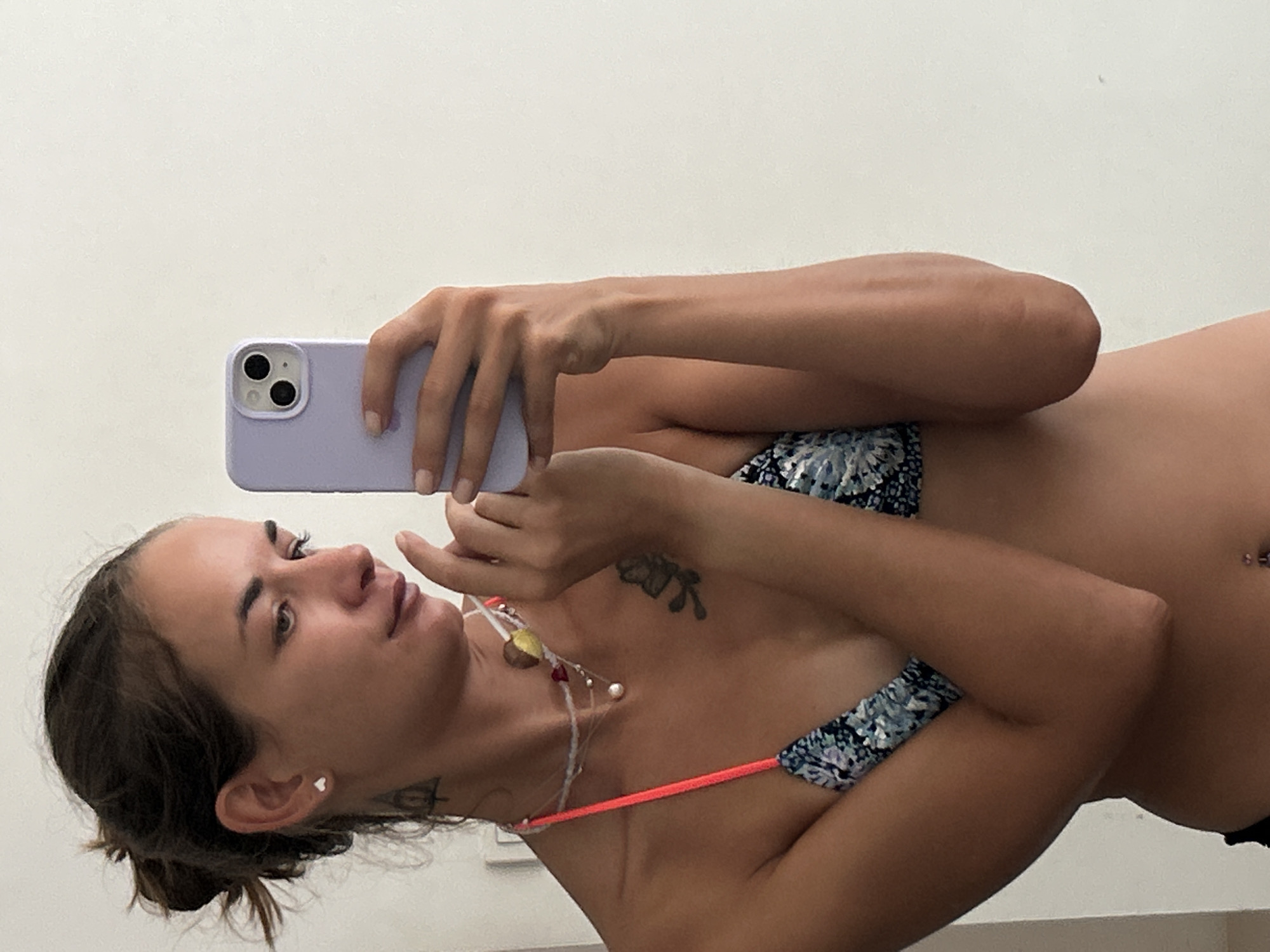 A woman is taking a mirror selfie indoors. She has her hair pulled back in a bun and is wearing a bikini top and a necklace. She is holding a light purple smartphone in her right hand and is looking at her reflection. Her left hand is slightly raised and resting on her chest. The background appears to be a plain wall. This image was taken in Kathu, Thailand.