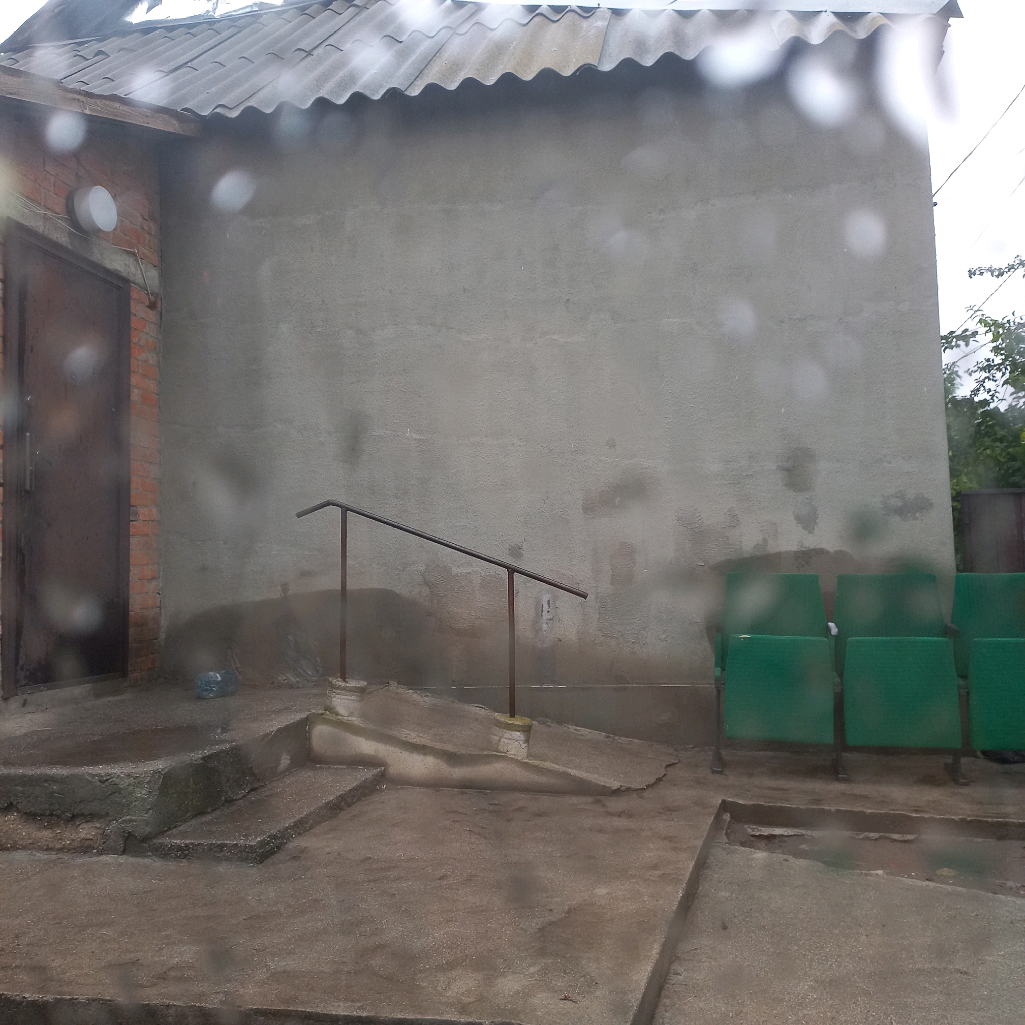 The image depicts an outdoor scene, possibly a back entrance or service area of a building, with a grey, textured wall as the dominant feature. To the left, a dark metal door is set into a brick facade. Above the door, a circular light fixture is mounted. A low, sloped ramp with a metal handrail leads up to a higher level. In the foreground, there are concrete steps and pathways. To the right, a row of green metal benches is visible. The ground appears wet and reflective, suggesting it has recently rained or is currently raining, with circular water droplets present on the lens. Some debris, including a blue plastic bottle, is scattered on the ground. The overall impression is one of a utilitarian, somewhat run-down urban environment.