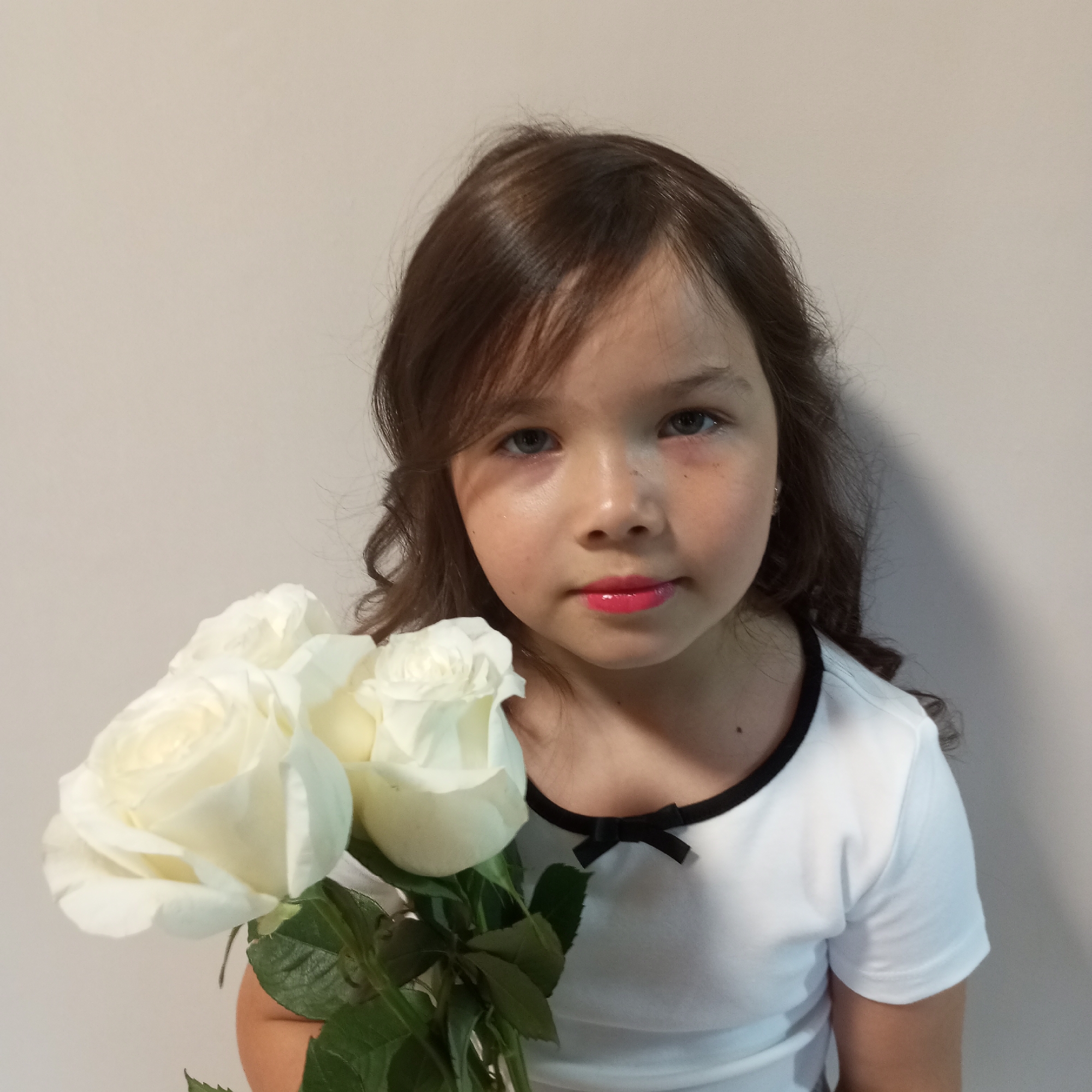 A young girl with long, dark brown hair and a light complexion holds a bouquet of three white roses. She is wearing a white t-shirt with a black trim around the neckline, which features a small black bow. The girl has bright blue eyes and pink lipstick. She is positioned in front of a plain, light-colored wall. The lighting suggests an indoor setting, possibly a room or a studio. There is no discernible urban environment or specific location within Pervomaysky, Russia indicated in the image. The image appears to be a portrait.