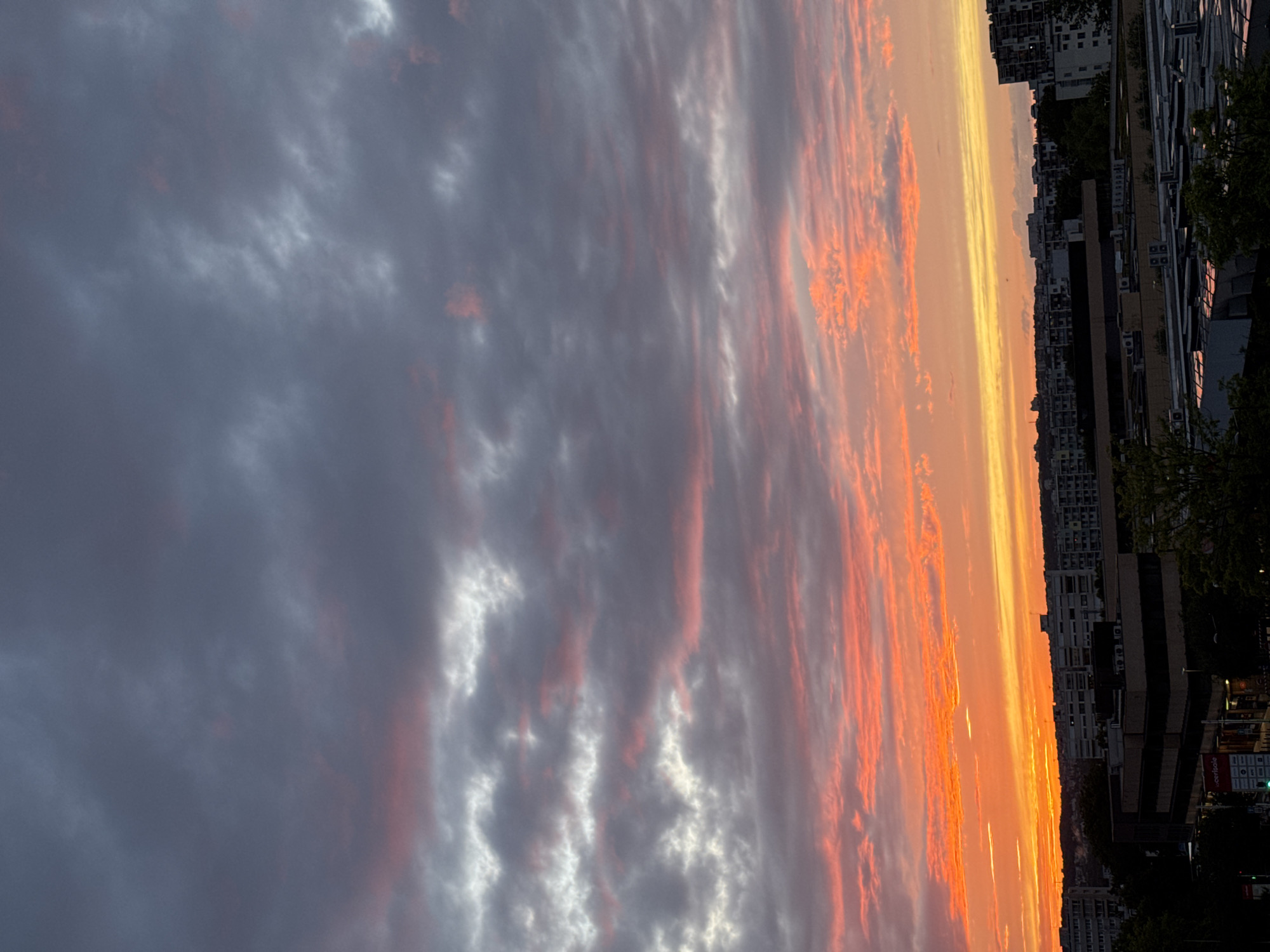 The image captures a dramatic sunset over the city of Fresnes, France. The sky is filled with a vibrant display of colors, transitioning from a bright orange and yellow horizon to streaks of pink and purple, eventually fading into dark gray clouds higher up. These clouds are textured and appear to be illuminated by the setting sun from below.
In the foreground, silhouetted against the colorful sky, are the rooftops of buildings in Fresnes. These appear to be primarily apartment buildings and other urban structures, with some trees visible between them. The buildings are dark, with only the faint outlines of their shapes discernible. The scene suggests an evening view from a high vantage point, likely an apartment or office building, looking out over the urban landscape as day turns into night. There are no people or active events visible in the image. The overall impression is one of natural beauty juxtaposed with the man-made environment of the city.
