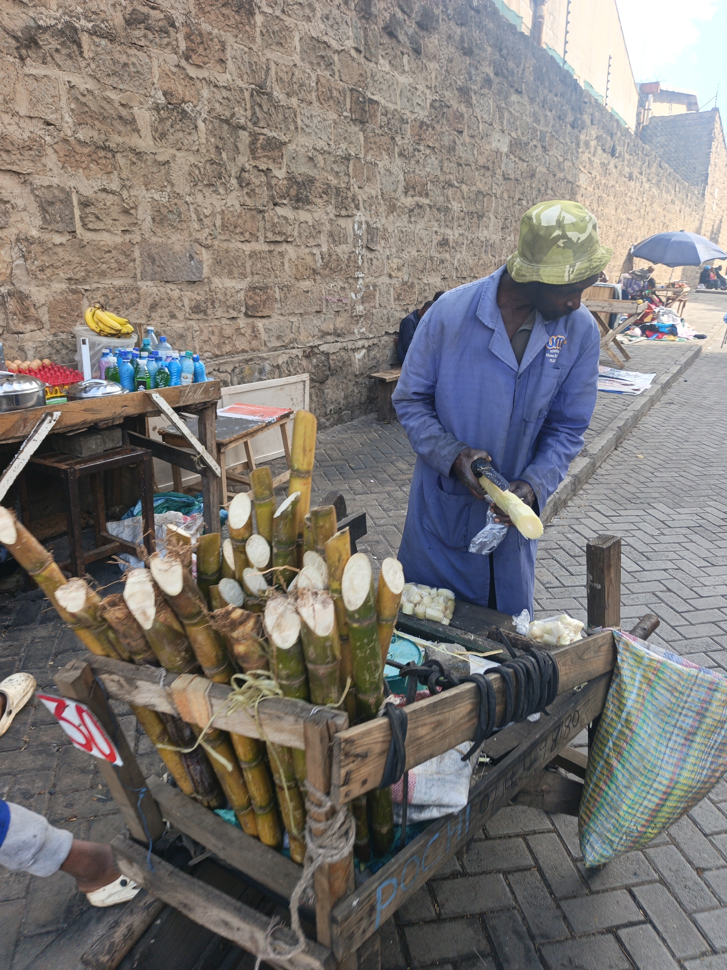 The image depicts a street vendor in Nairobi, Kenya, selling sugar cane. The vendor, wearing a blue work coat and camouflage bucket hat, is peeling a section of sugar cane. He is standing behind a wooden cart filled with stalks of sugar cane that have been cut into sections. The sections are bundled together with string. The cart appears to be crudely constructed. To the left of the sugar cane cart, there is a makeshift table displaying fruits, drinks and eggs. The background is a high stone wall. The street is brick-paved. Other people are visible in the background under an umbrella.