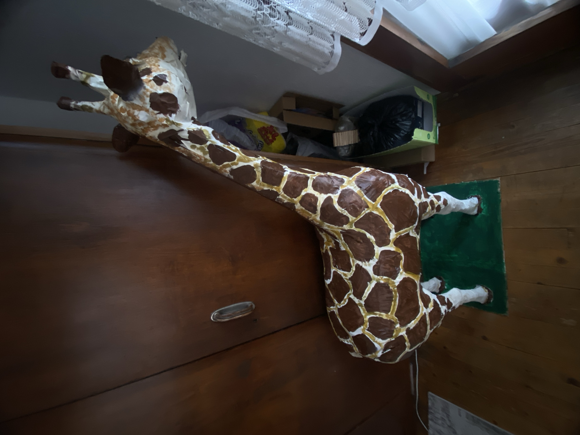 A life-sized giraffe statue lies horizontally atop a wooden surface, likely a cupboard, in an indoor setting. The giraffe is painted with white and brown spots, typical of the animal's natural coloration. The head and neck extend towards the left side of the frame, while the body rests on a green mat, placed on the right side. The statue appears to be somewhat aged or handmade. The environment consists of wooden walls and a cluttered shelf above the giraffe, containing various objects. The exact location in the city of Blagoevgrad, Bulgaria is not ascertainable from the given image.