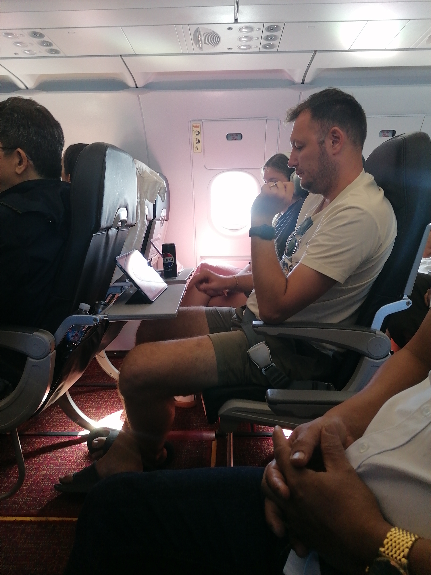 The image shows the interior of a passenger airplane cabin. A fair-skinned man with short hair is seated, wearing a light-colored t-shirt and shorts. He has a seatbelt fastened and is looking towards the left of the frame, resting his chin on his hand. Sunglasses are hooked onto his shirt. A woman is seated next to the window, behind him. A tablet is placed on the tray table in front of him, alongside a can of Pepsi. To the left, a person is seen wearing a black jacket. To the right, partial view of a person seated wearing a white shirt and a gold watch. The carpet is red. The lighting suggests it is daytime. The location is Unknown, Unknown.
