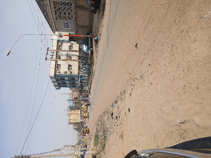 The image displays a street scene in Mgbidi, Nigeria, captured during daylight under a clear, pale blue sky. The foreground features an unpaved, dusty earthen shoulder on the left, partially revealing the front left fender and headlight of a dark-colored vehicle. The roadway transitions into a central paved section, flanked by unpaved, debris-strewn shoulders.

On the left side of the paved road, three yellow auto-rickshaws (Keke Napep) are visible, oriented away from the camera. Multiple pedestrians are observed near these vehicles, with one individual appearing to interact with an auto-rickshaw. Utility poles, some wooden and others concrete, support a dense network of overhead electrical wires crisscrossing the sky.

On the right side of the street, a multi-story building with a beige facade and prominent blue trim, featuring multiple exterior air conditioning units, stands behind a transformer structure. Adjacent to this building is another structure with a grey tiled roof, a grey concrete perimeter wall, and windows fitted with decorative security bars, one of which displays "NNN." A modern streetlight with a tall pole and an orange-red base is situated in front of this wall. Further along the right shoulder, a blue-green sedan and a dark-colored sedan are parked, both facing away from the viewer.

Numerous additional buildings, varying in height and construction, line both sides of the street in the midground and background, indicating a developed urban area. Scattered litter and tire tracks are present on the unpaved sections of the road.