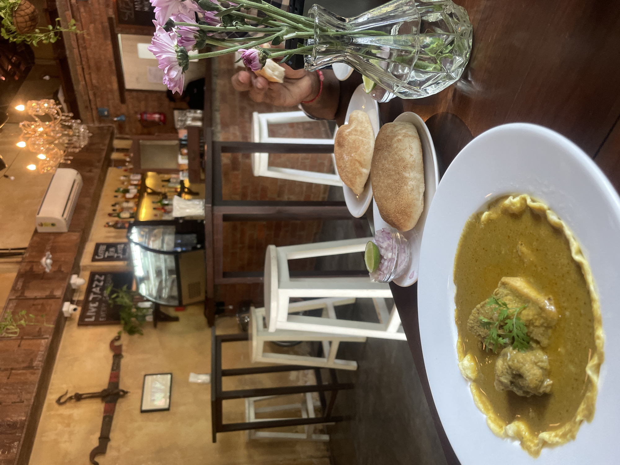 This image captures an indoor dining scene at a restaurant or cafe, likely in Chopdem, India. The foreground features a meal set on a dark wooden table. A large white bowl contains a vibrant yellow-green curry, with pieces of meat and fresh green herbs (likely cilantro) as a garnish. Alongside, a small white plate holds two light-colored bread rolls, and a tiny container offers chopped red onions and a lime wedge. A clear, cut-glass vase holds a bouquet of light purple chrysanthemums, adding a touch of elegance to the table. A person's hand is visible near the vase, holding a piece of bread, suggesting the meal is about to be enjoyed or is already in progress.
The background reveals the cozy and rustic interior of the establishment. Exposed brick walls and wooden beams create a warm ambiance. A bar area is visible with bottles of alcohol and a display fridge. White and dark high stools are arranged along a counter. Overhead, a sparkling chandelier provides additional lighting, and a white air conditioning unit is mounted on the wall. Decorative elements include a large, old-fashioned balance scale hanging on a textured wall, and a blackboard sign that reads "LIVE JAZZ," indicating entertainment is offered. The overall setting suggests a casual yet inviting atmosphere for dining.