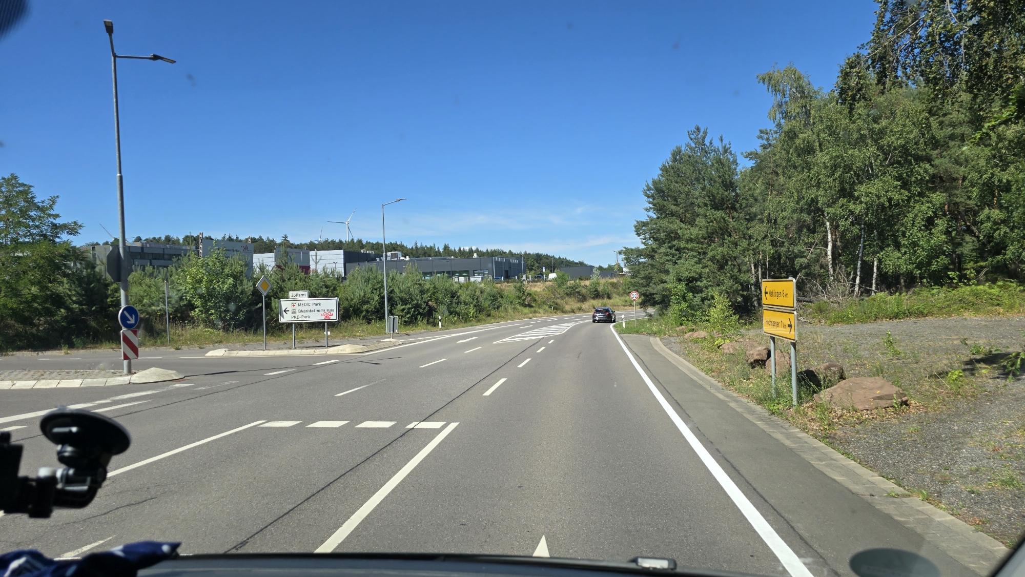 The image shows a two-lane road in Kaiserslautern, Germany, on a clear day with a blue sky. The road has white lane markings and is bordered by trees and vegetation. In the foreground, a car's dashboard and windshield are visible, along with a suction cup mount for a device. To the left of the road, there is a street lamp, a traffic sign with a blue arrow pointing down, and a red and white striped pole. Further down the road, a white sign indicates "ZOLLAMT", "MEDIC Park", "Erlenbad", and "PRE-Park". A black car is visible driving on the right lane of the road. To the right of the road, there is a yellow directional sign pointing left to "Mörlbach 8 Km" and right to "Hochspeyer 7 Km". Trees and rocks line the right side of the road. In the background, there are buildings and wind turbines.