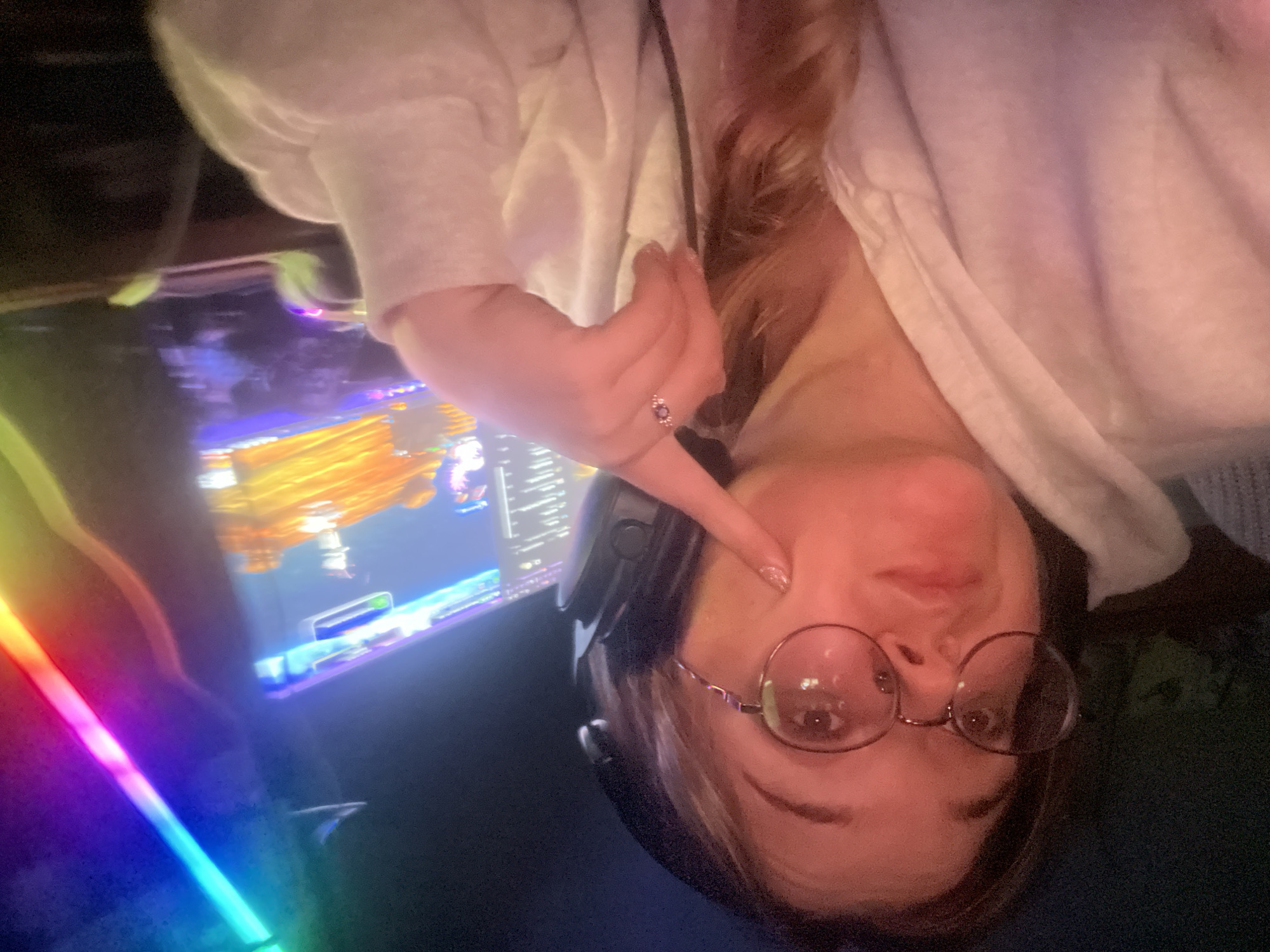 The image shows a young woman wearing glasses and headphones, with her right index finger touching her cheek. She is wearing a white hoodie and a ring on her left hand. Behind her, a computer screen displays a game with bright, colorful graphics, and RGB lighting is visible on the right side of the frame. The overall scene suggests a gaming or entertainment setting. There is no indication of the location being in Kolomna, Russia.