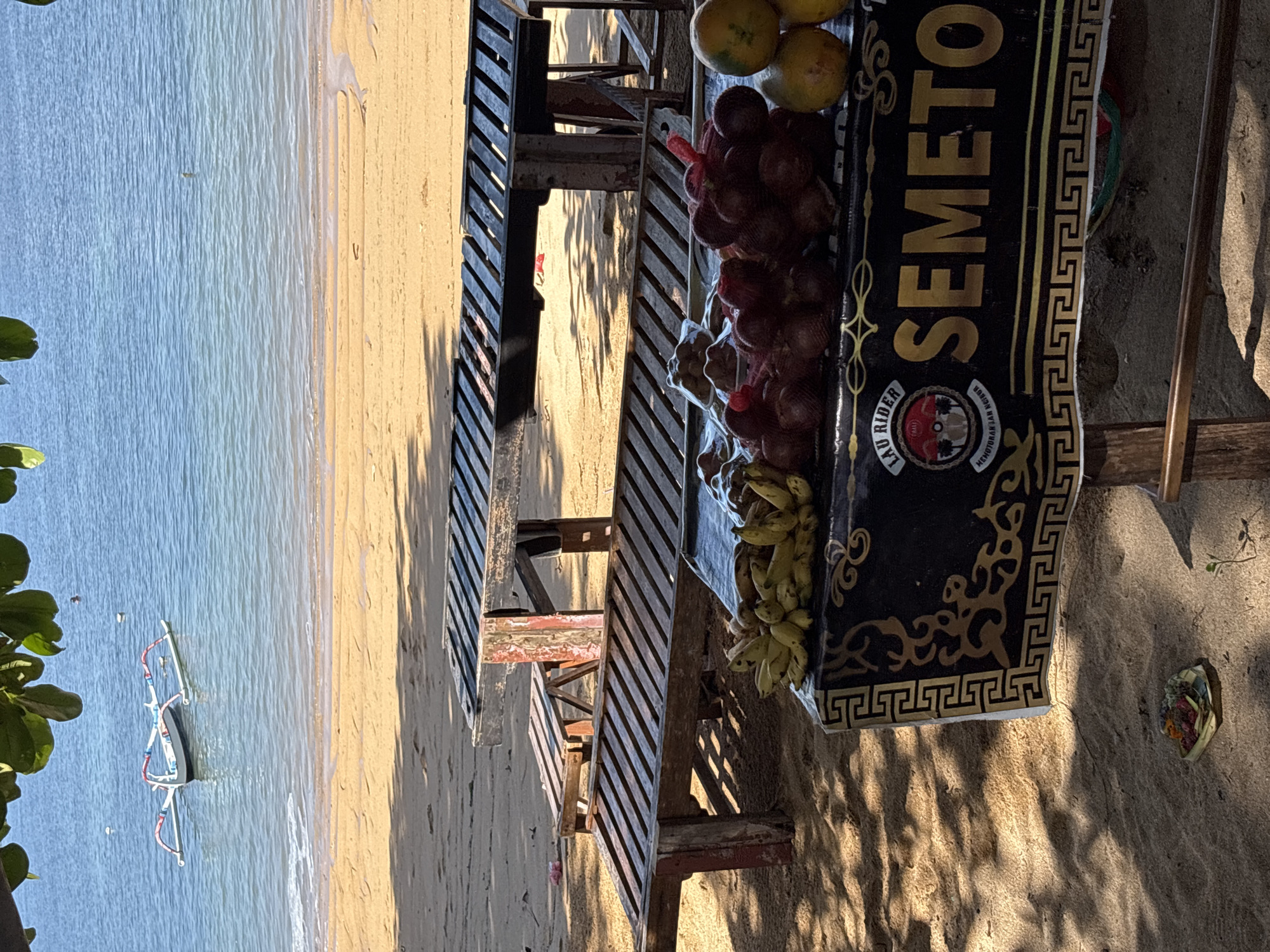The image depicts a beachside scene, possibly in or near Denpasar, Indonesia. A wooden table laden with fruit, including mangoes, bananas, and several bundles of what appear to be red salak fruit, is a central element. The table has a black sign with gold lettering that reads "SEMETON". A simple wooden structure, possibly a beach stall or table, supports the fruit. To the left, there's a view of the beach, with gentle waves and a small boat in the distance. The background shows sand and some shadows, suggesting the scene is photographed during the day. The general atmosphere conveys a beach vendor setting.