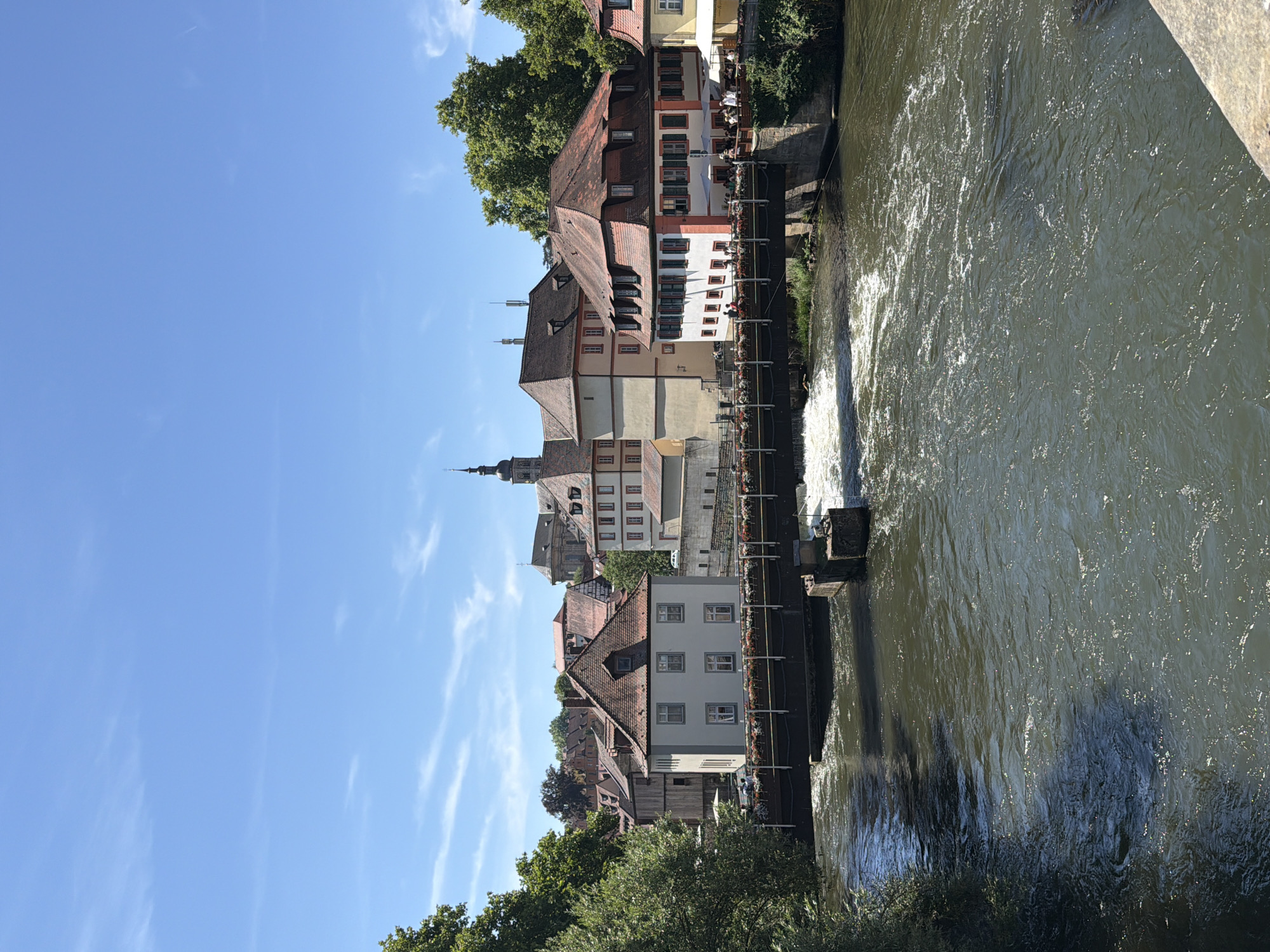 This is a landscape photograph of the Regnitz River in Bamberg, Germany, taken on a sunny day. The river flows from the foreground towards the background, with a bridge spanning across it. Along the riverbanks, there are several buildings with red tiled roofs, typical of Bamberg's historic architecture. Some of these buildings appear to be restaurants or cafes, with outdoor seating visible on the bridge and along the bank. Trees line the banks of the river, with lush green foliage. The sky is blue with a few wispy clouds. The scene depicts a serene and picturesque urban environment.
