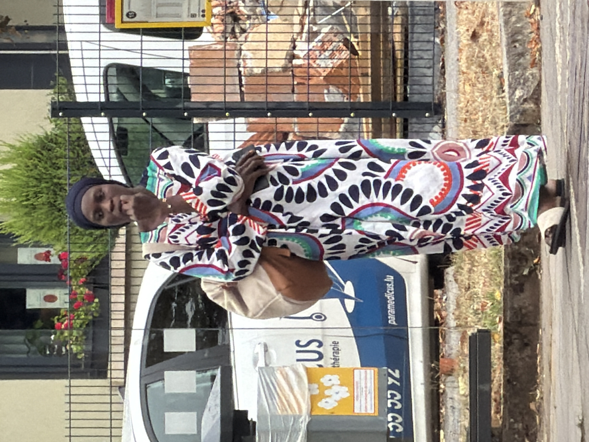 A Black woman wearing a colorful, patterned long dress and a dark headscarf stands on a sidewalk in front of a parked white van with "paramedicus.lu" written on its side. The van is branded with medical symbols and text, suggesting it is an ambulance or medical service vehicle. The woman is holding a tan bag on her left shoulder and appears to be bringing her left hand to her mouth. She is wearing sandals. Behind the van, a black metal fence separates the sidewalk from a pile of red bricks and construction debris. In the background, a building with windows displaying flowers can be seen. The scene appears to be in a city environment, possibly in Esch-sur-Alzette, Luxembourg, given the ".lu" domain on the van.