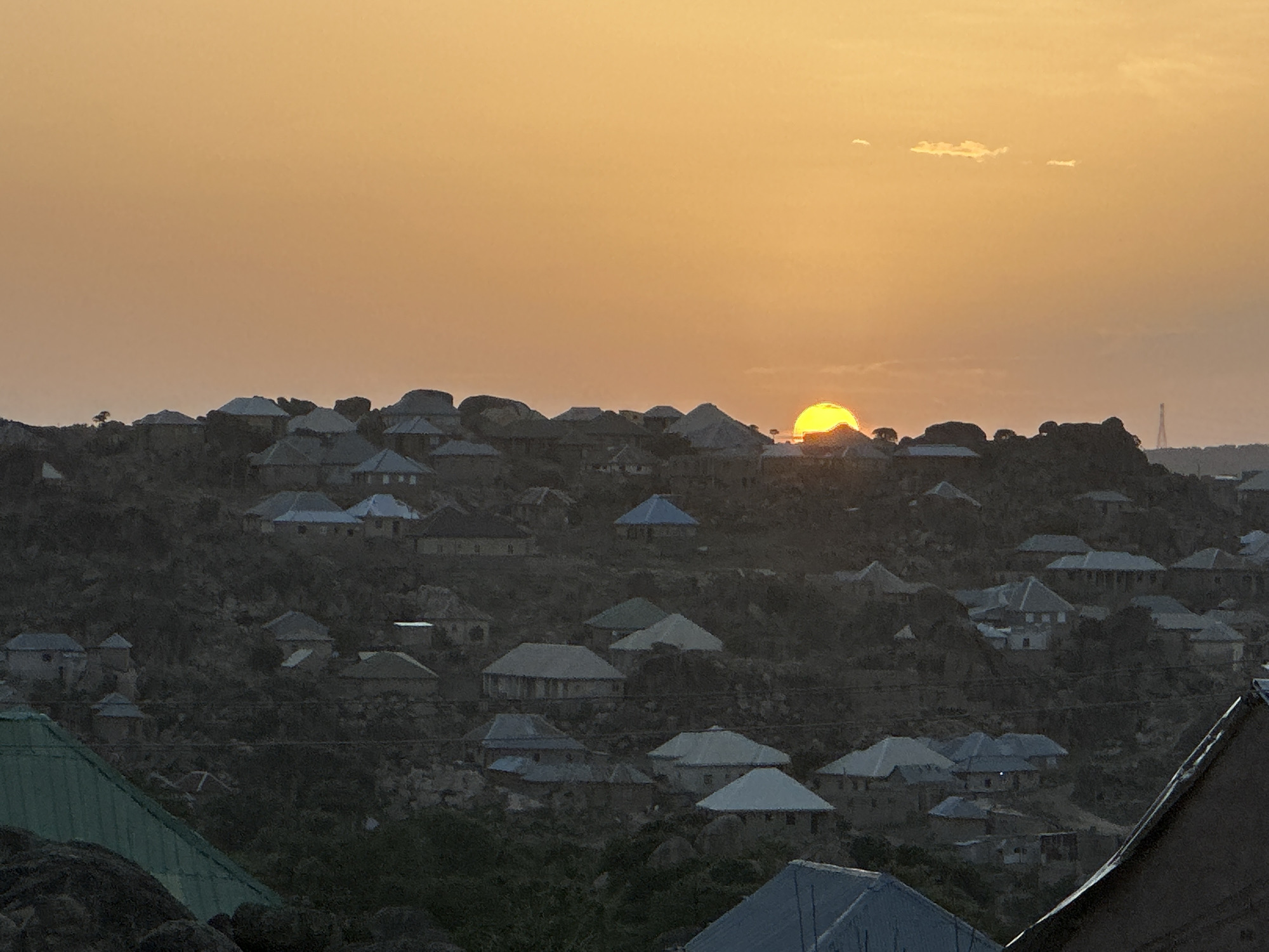 The image depicts a sunset view of a village scene in Jos, Nigeria. The sun is partially obscured by a hill covered with closely situated houses, most with light-colored roofs. The sky is a gradient of orange hues. There is a distant telecommunications tower on the right of the image. The foreground includes additional rooftops and rocky terrain.