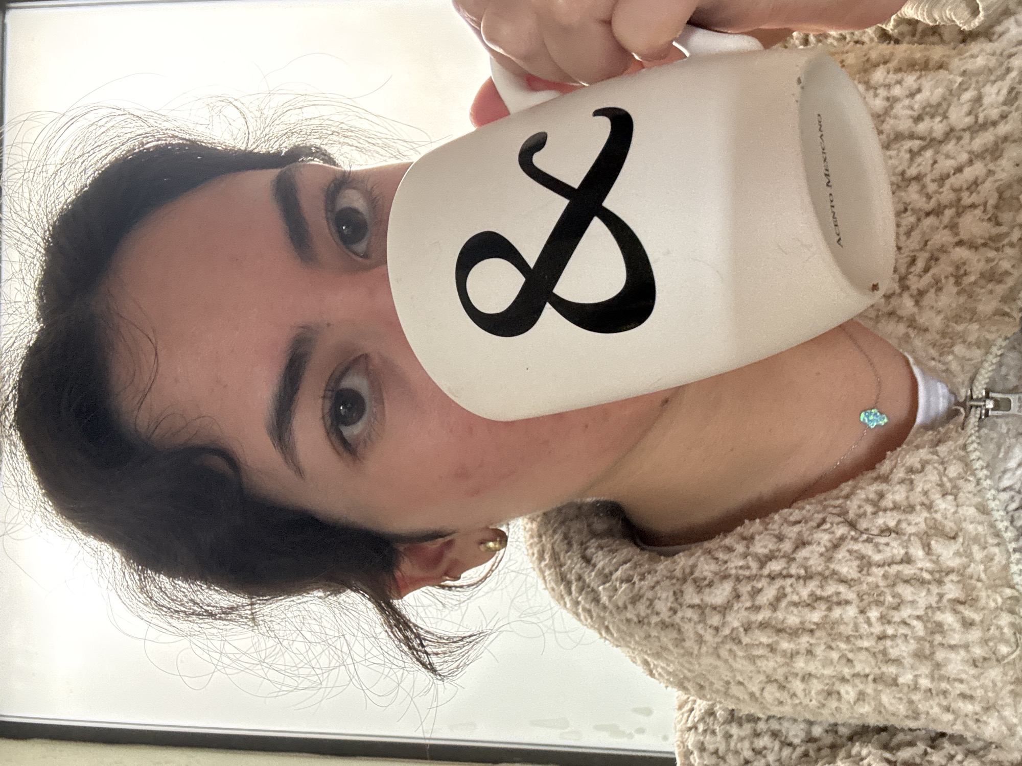 A young woman is holding a white mug with a black infinity symbol. The mug has the text "ACENTO MEXICANO" in small font on its side. The woman has dark hair and light skin. She is wearing a beige, fluffy sweater, and a silver necklace with a small, blue, cloud-shaped pendant. The location is not specified or visible in the image.