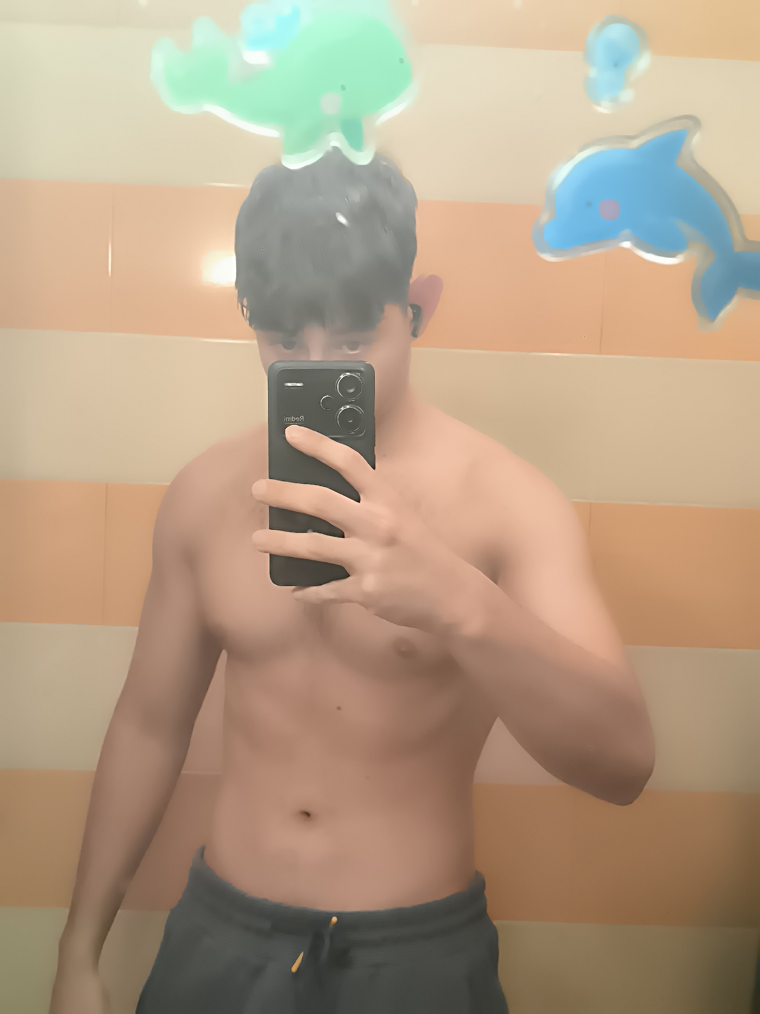 A shirtless person is holding a smartphone, taking a picture of themself in a bathroom. The background is an orange-and-cream striped wall with decorative plastic dolphins and sea creatures. The person is wearing dark-colored sweatpants with a drawstring. The smartphone is black and has two visible cameras. The location is unknown, but the city is mentioned as Sofia, Bulgaria. However, no features of Sofia are apparent in the image.