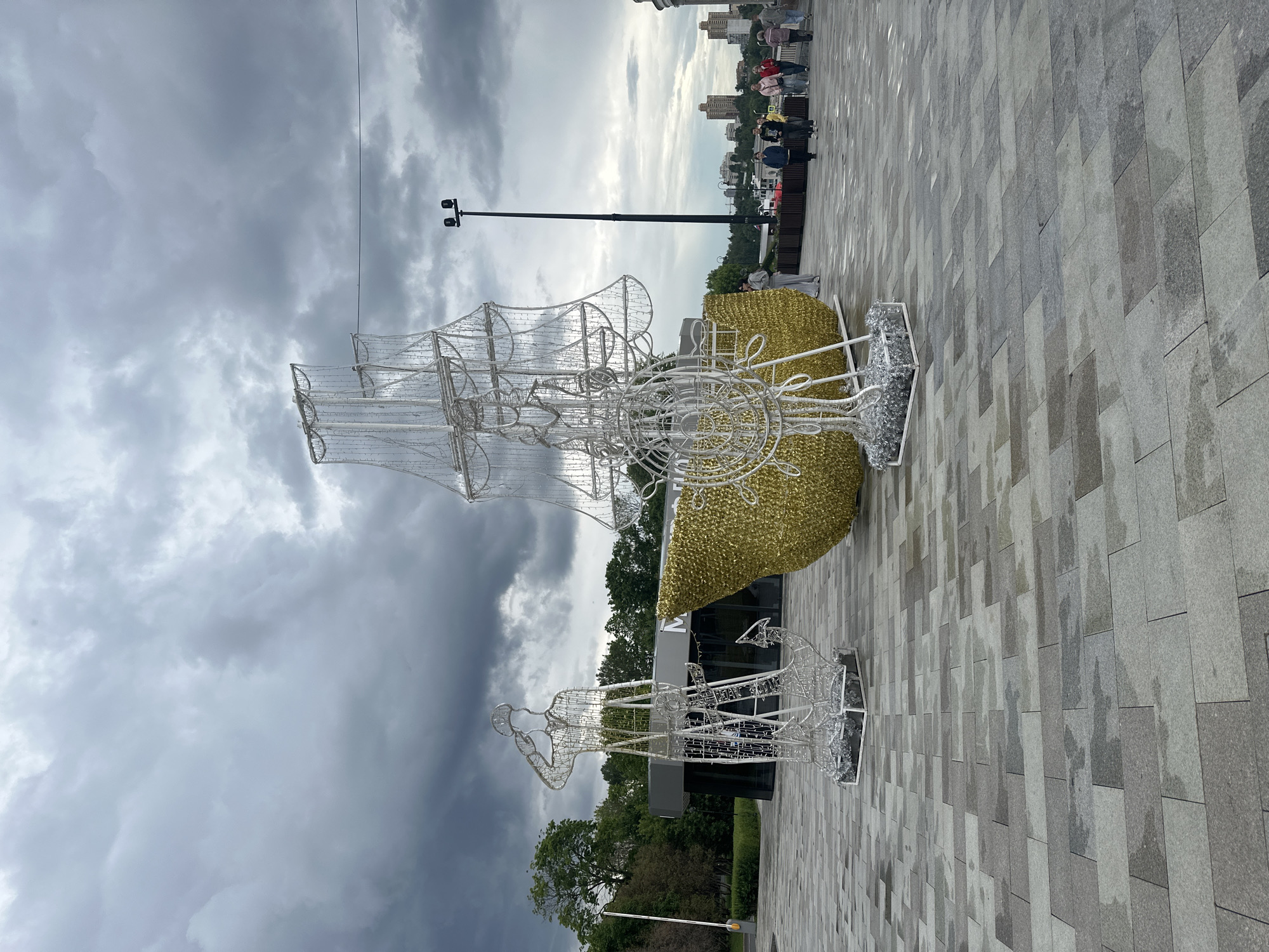 An outdoor, wide shot shows two illuminated wire sculptures of a ship and a sailor on a paved area. The sailor sculpture is on the left, saluting, with an anchor at its base. To its right is a large, golden ship hull with a wireframe superstructure and sails. The sculptures are in a public square with trees and a modern building in the background. People are walking on a walkway behind the sculptures, with buildings visible in the distance. The sky is overcast with dramatic clouds.
