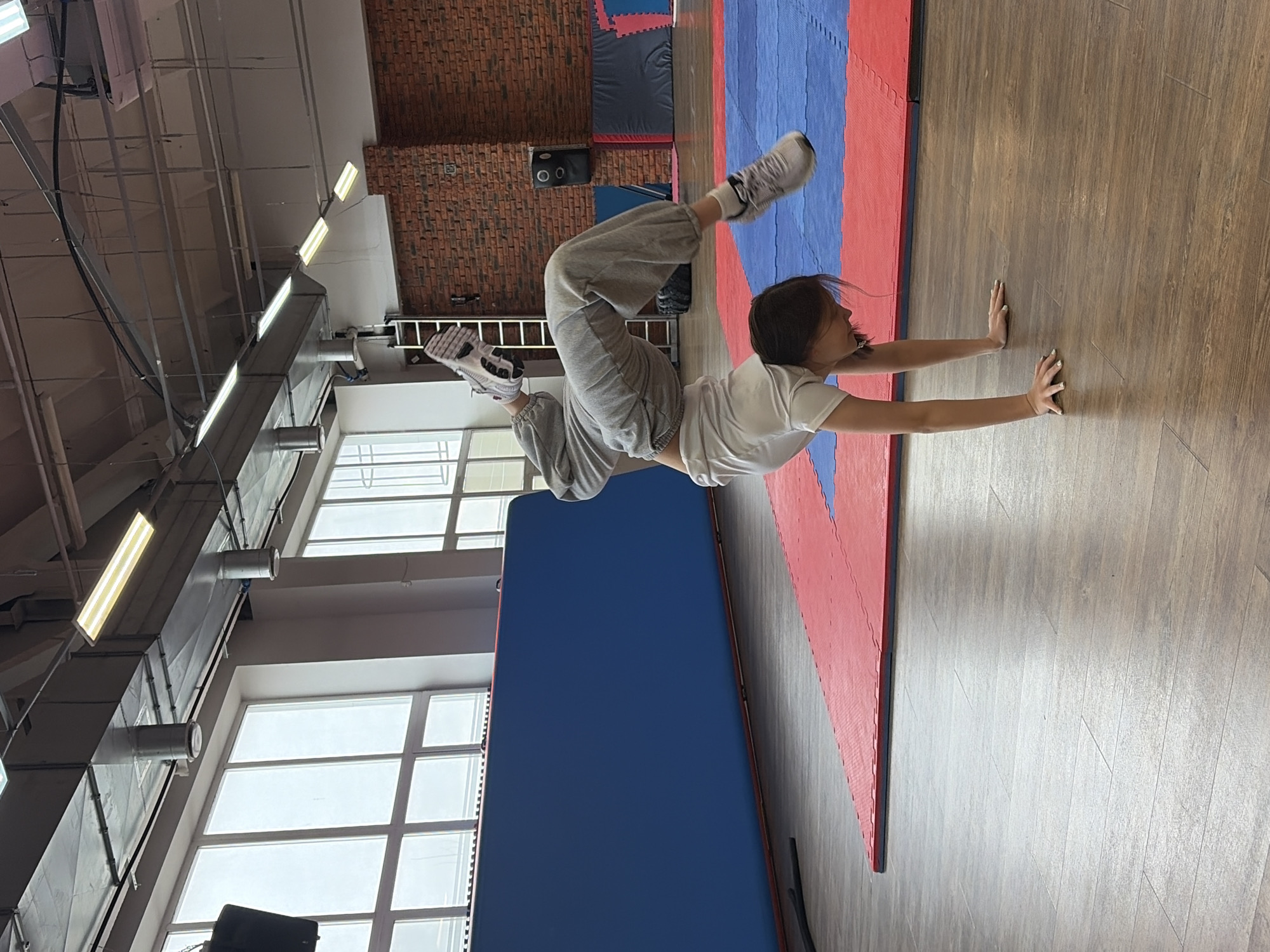 A young woman is practicing handstand push-ups in what appears to be a gym or martial arts studio. She is wearing a white t-shirt and gray sweatpants, with gray sneakers on her feet. Her hands are placed flat on the wooden floor, supporting her weight, while her body is inverted with her legs pointing upwards. The gym has interlocking mats in red and blue along the wall and a brick wall is on the other side of the room. There are windows and ventilation ducts and lighting on the ceiling. The location is Chaikovsky, Russia.