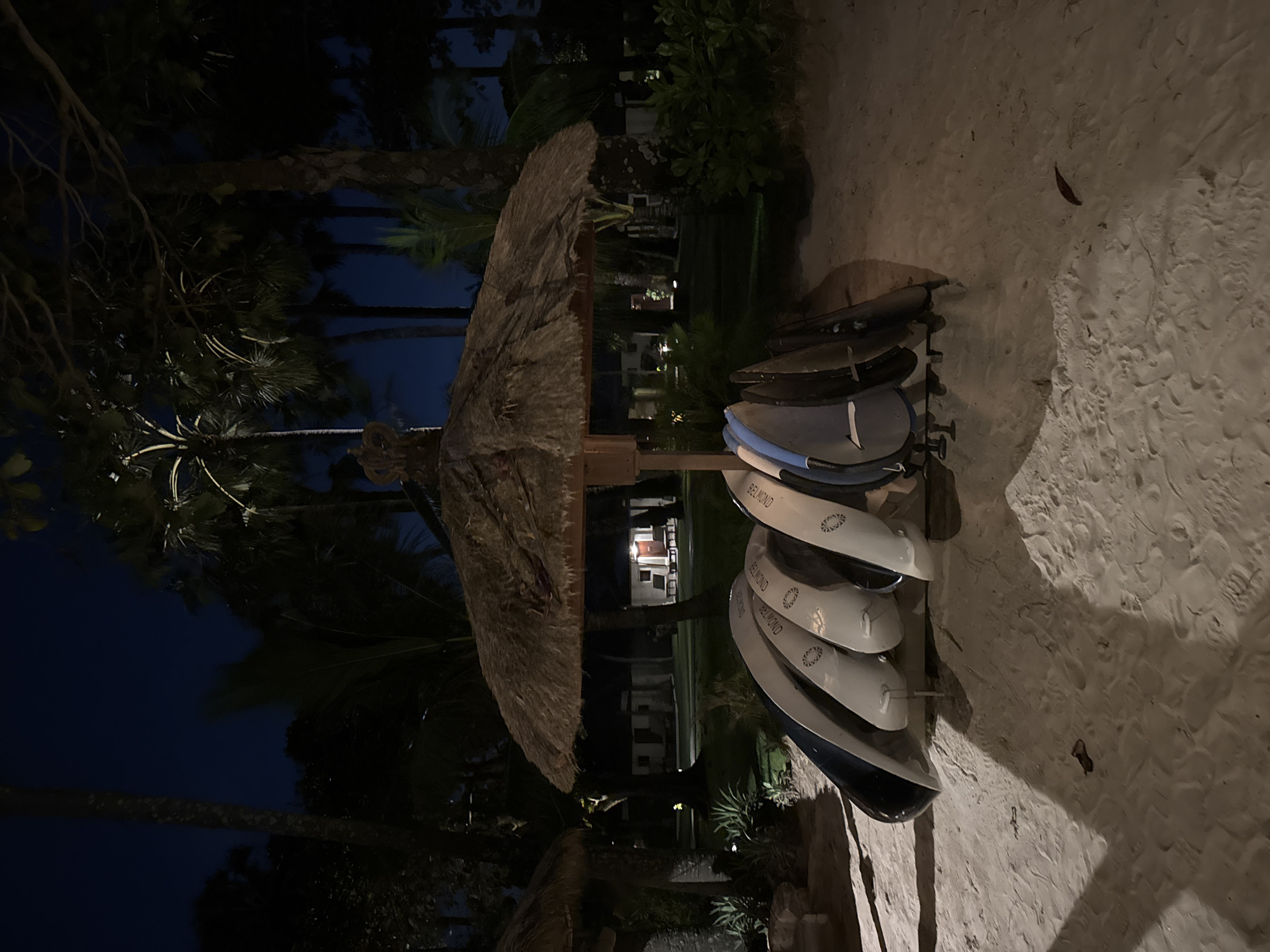 This nighttime image depicts a tranquil resort or beach club setting, consistent with a tropical location like Kuta Selatan, Indonesia. The scene is dominated by a deep indigo sky overhead, with numerous palm trees and other tropical foliage silhouetted against it, their dark forms suggesting lush surroundings.
In the mid-ground, a large, circular thatched umbrella stands prominently, its texture visible under soft artificial lighting. To its right, a rack holds several stand-up paddleboards or surfboards. These boards are stacked horizontally, and on each visible one, the word "BELMOND" is clearly legible in white text, accompanied by a circular logo. This branding indicates the location is associated with the luxury Belmond hotel group.
The ground consists of light-colored sand, showing faint impressions and textures, with a single dark leaf resting on its surface. In the background, partially obscured by trees, some dimly lit buildings or structures are visible, suggesting hotel accommodations or resort facilities. No people are present, and no specific activity is occurring, contributing to a peaceful and serene atmosphere. The image captures the quiet beauty of a tropical evening, with recreational equipment awaiting the next day's activities.
