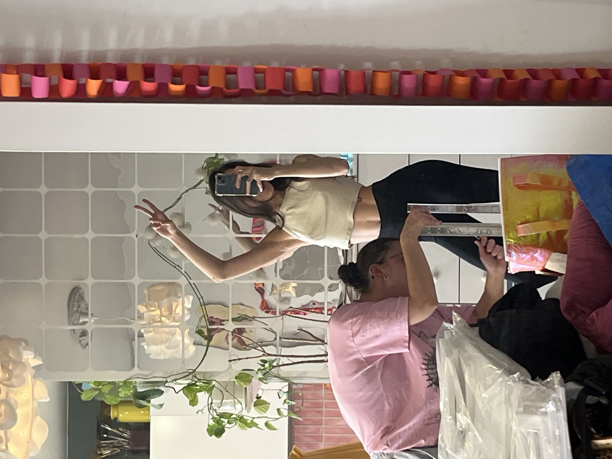 The image depicts a reflection in a mirror of two women and their surroundings. The reflection also shows paper chain decorations hanging above the mirror.
One woman is holding a phone and appearing to take a photo, and the other is standing next to her.
The details of the location within the city of Krasnoyarsk, Russia, cannot be determined.