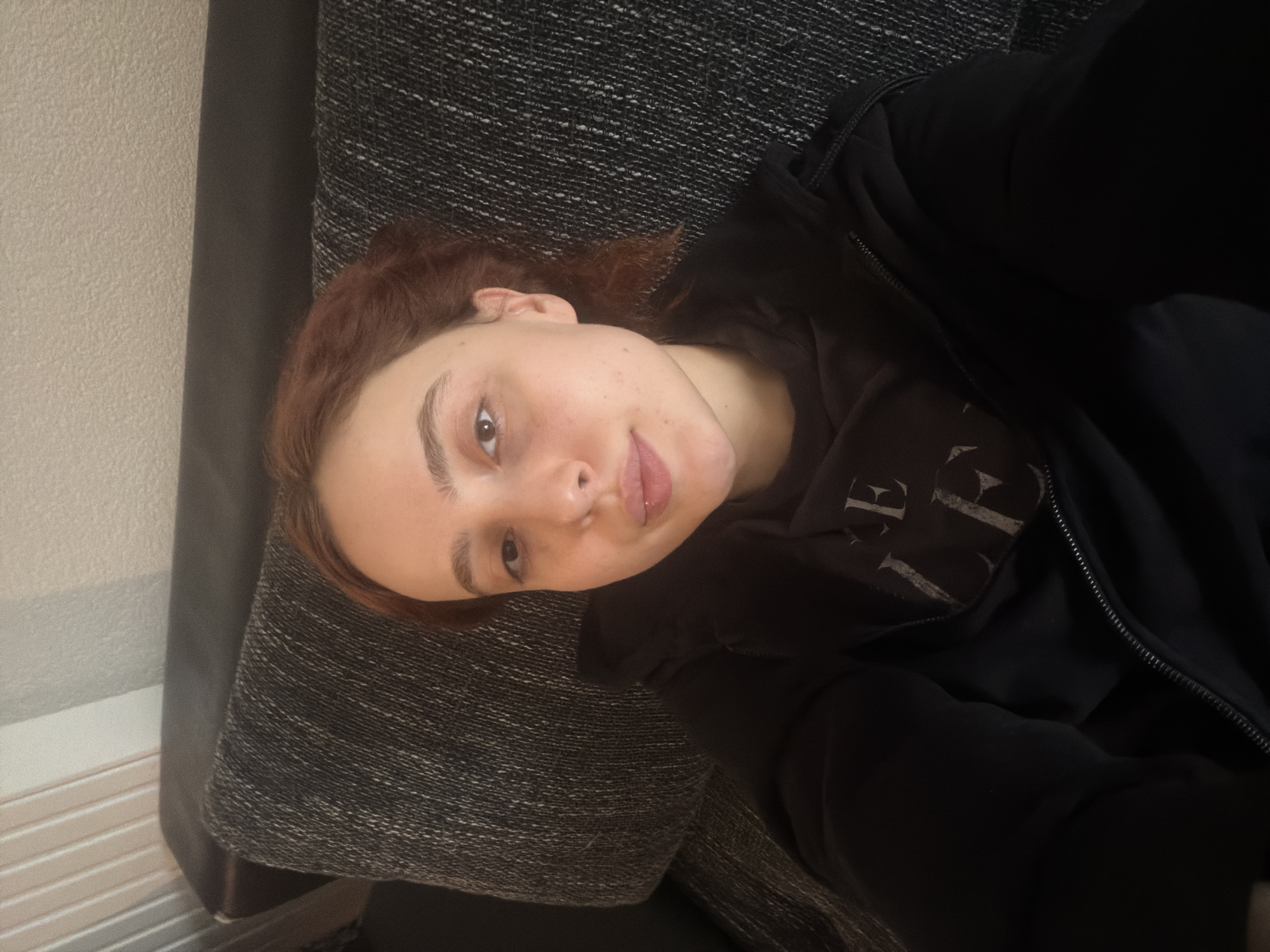 A young adult female subject with fair skin, light brown to auburn hair, and brown eyes is depicted in a medium-shot, upward-angled photograph. She is looking directly at the camera with a neutral expression, her lips slightly parted. The subject wears a dark, possibly black, zip-up hooded sweatshirt over a dark t-shirt, which displays partial light-colored text, appearing as "CE LE". She is reclined against a dark grey, textured fabric backrest of a sofa or couch. A darker, plain headrest is visible behind her upper head. The background consists of a light-colored wall with vertical paneling on the upper left, potentially a radiator cover, and a textured, off-white wall above the furniture. The setting is an indoor residential environment. No specific visual markers are present in the image to determine the exact location within Strasbourg, France.