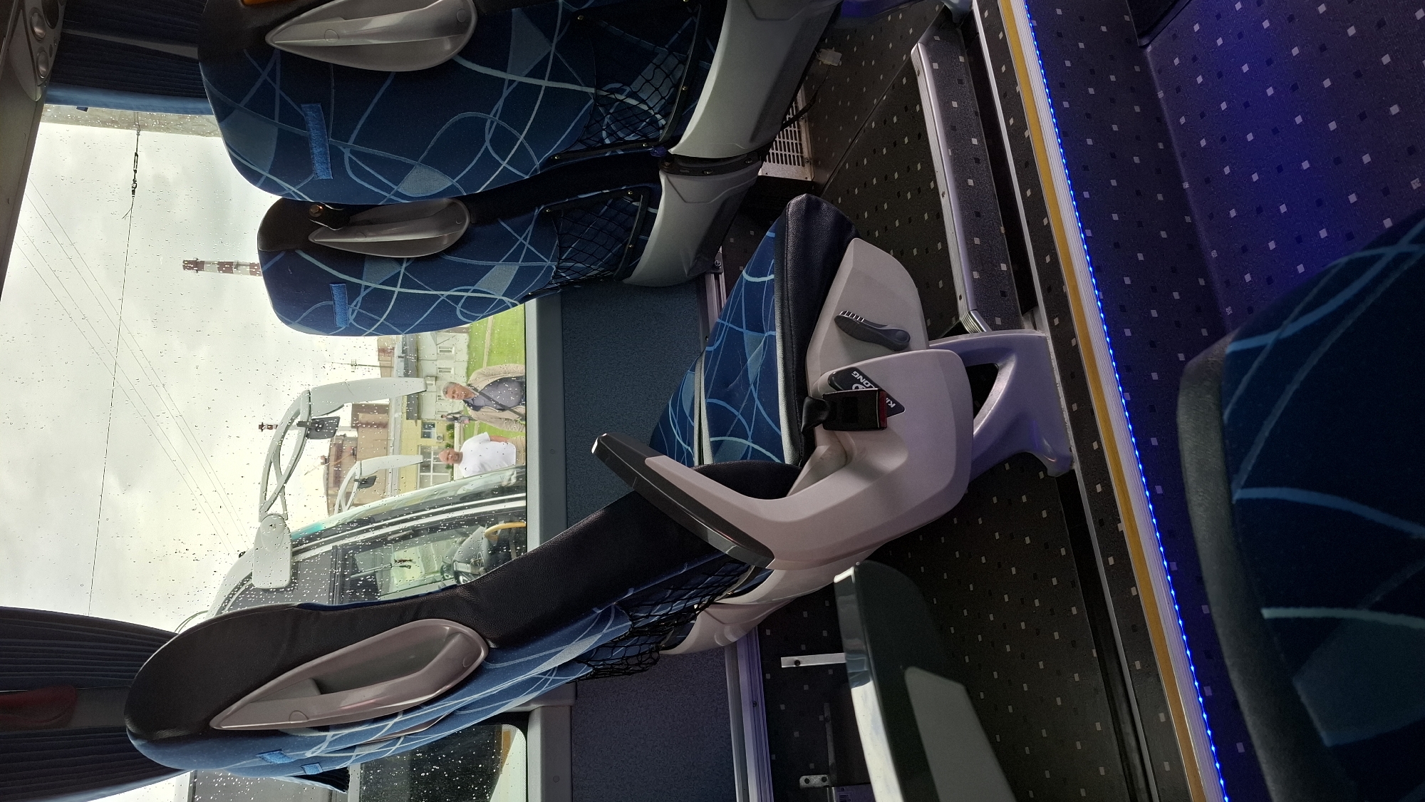 The image shows the interior of a bus, likely on a rainy day, as evidenced by water droplets on the window. The seats are upholstered in blue fabric with a geometric pattern. One seat is in the foreground, a lighter gray color with a black seat cushion, and appears to be a pilot's or driver's seat due to its central position and armrest configuration. There is a black object resembling a shoe placed on the armrest of this seat. Text visible on a label on the seat states "KING LONG".
Through the window, two men are visible standing outside the bus. They appear to be engaged in conversation. The weather seems overcast. In the background, there are buildings and a tall industrial structure, possibly a smokestack. The bus interior is illuminated by blue LED lights running along the aisle. The overall scene suggests a stop or a pause in the bus journey, possibly at a service area or a point of interest in Natal'ino, Russia.