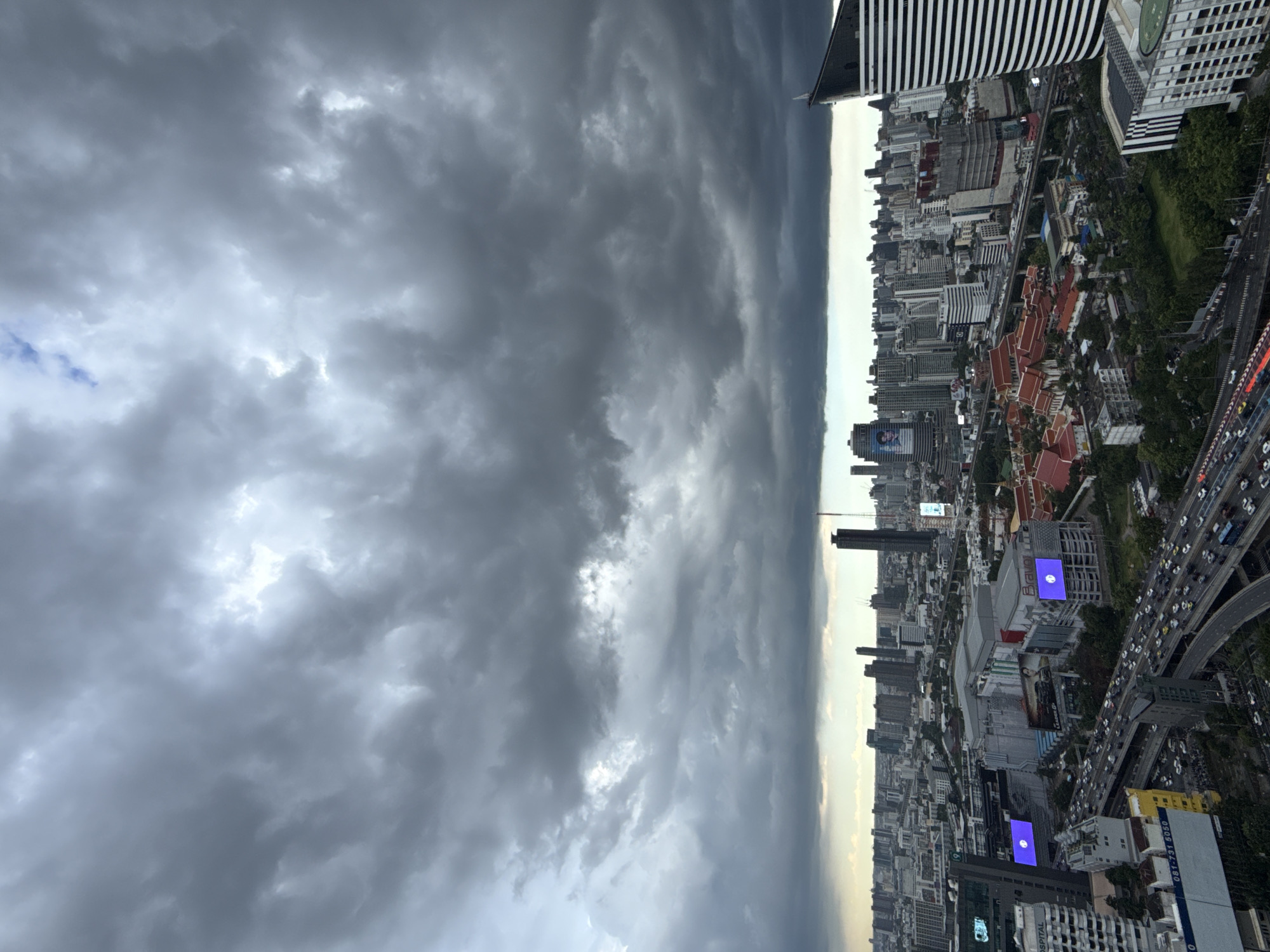This media file captures a dramatic, overcast sky over a dense urban landscape, likely Bangkok, Thailand, given the provided context. The image is oriented vertically, with the sky dominating the left half and the cityscape filling the right.
The sky is filled with heavy, dark, and textured clouds, suggesting an impending storm or recent rain. Pockets of brighter light break through in the upper left, hinting at the sun struggling to penetrate the thick cloud cover.
Below the sky, a sprawling city unfolds. Numerous tall buildings, ranging from modern skyscrapers with glass facades to older structures with distinctive tiled roofs, are densely packed together. Greenery is visible in patches, particularly along what appears to be a major elevated highway or road system snaking through the city. Cars are visible on this roadway, suggesting it's a busy thoroughfare.
Notable details include two prominent bright blue rectangular digital billboards visible on buildings within the cityscape. One is situated on a lighter-colored building, and another on a darker one. Some text is visible on a blue and white sign in the lower right corner of the image, reading "081-731-5050". A large billboard featuring a person's face is also visible on a building in the middle distance.
The overall mood is one of a dramatic and possibly oppressive atmosphere due to the heavy clouds. The time of day is difficult to pinpoint precisely, but the subdued light suggests it could be late afternoon or early evening, or simply a heavily clouded day. The scene depicts the everyday hustle and bustle of a major city contrasted with the powerful presence of nature in the form of an imposing sky.