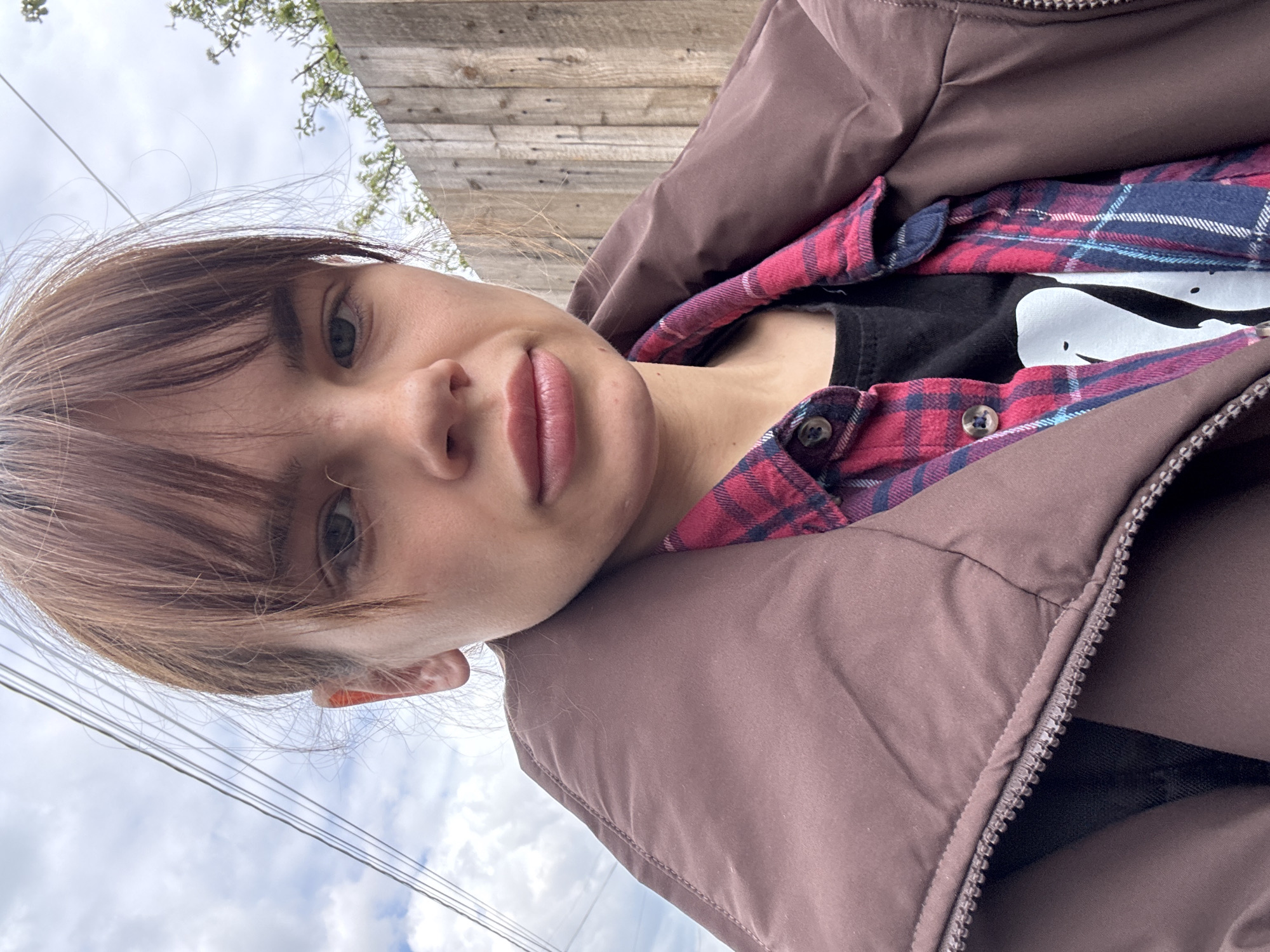 The image depicts a person with fair skin, brown hair with bangs, and light eyes. They are wearing a brown puffer jacket over a red and blue plaid shirt, which is layered over a black t-shirt with a white graphic. The person is likely outdoors, as a wooden fence, electrical wires, and a cloudy sky are visible in the background. The image appears to be taken at a low angle, looking up at the subject. There is no indication of the location being Rostov-on-Don, Russia.