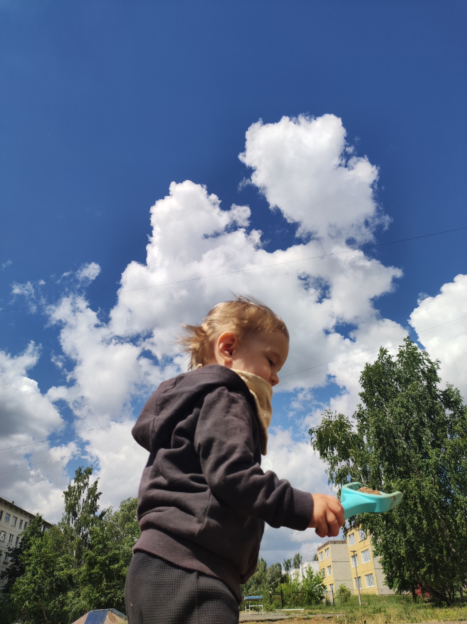 A low-angle, eye-level shot depicts a toddler in a brown hooded sweatshirt and dark pants, holding a blue toy shovel filled with dirt. The child is positioned slightly to the left of the center, looking down towards the right. The sky forms the backdrop, filled with white cumulus clouds against a blue sky. In the background, there are buildings with white walls and windows, and trees with green leaves. The scene suggests an urban park or playground setting. The location appears to be Chaikovsky, Russia based on architectural styles and vegetation. Electrical wires run across the sky near the cloud formations.