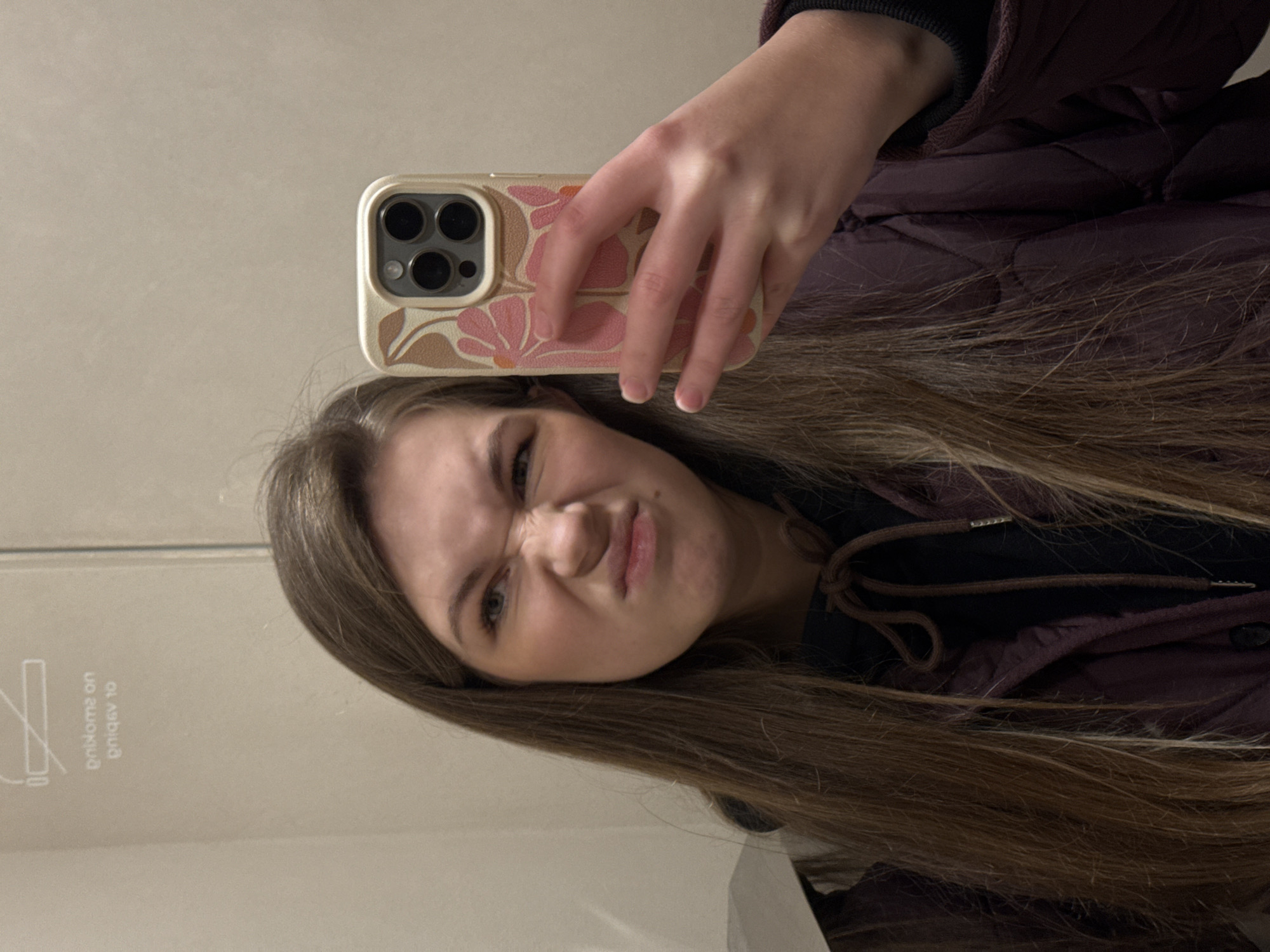 A young woman takes a selfie in front of a mirror. The woman has long brown hair and wears a dark jacket. She holds a smartphone with a floral case in her hand. The mirror shows a "no smoking" sign. It is not possible to determine the location in Belgrade, Serbia, from the given image.