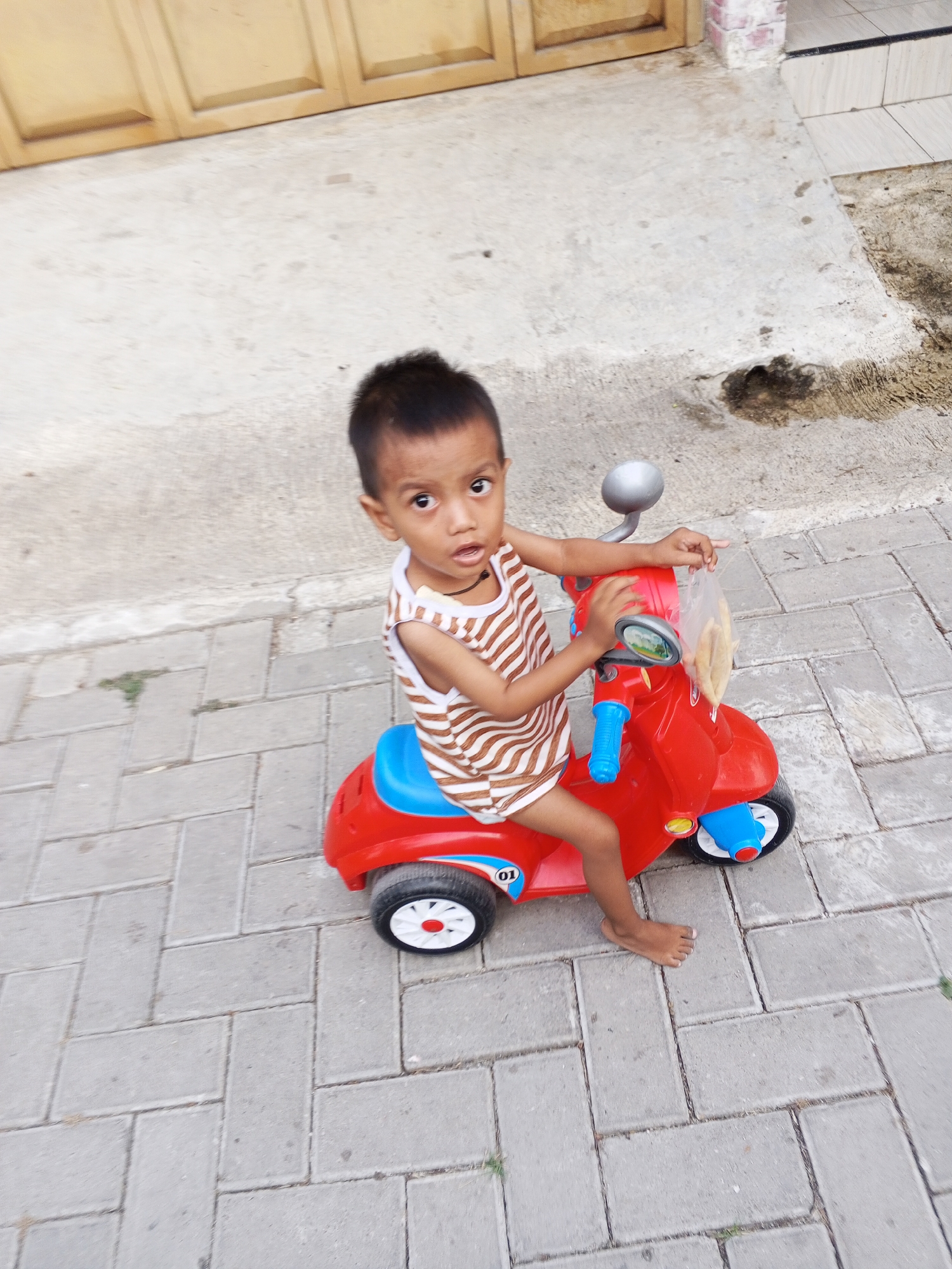 A young boy sits on a red toy scooter, his legs dangling over the side, on a paved walkway. He is wearing a sleeveless, horizontally striped shirt. The scooter has blue handlebars and accents. In the background, a beige-colored wall and part of a door are visible. The walkway is made of grey interlocking pavers. The scene appears to be outdoors, possibly in a residential area in Ciruas, Indonesia.