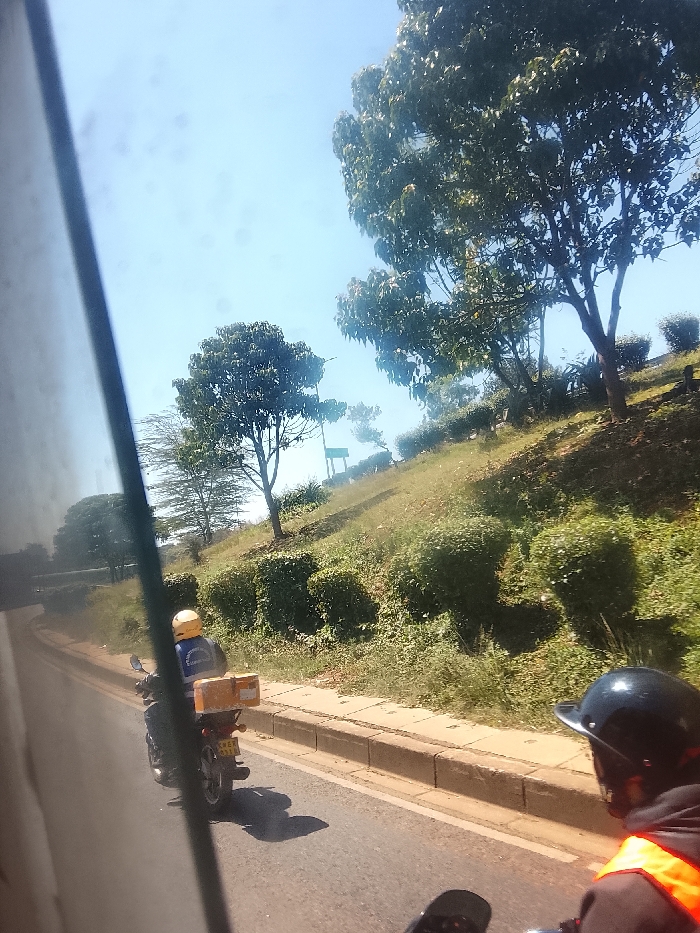A view from a vehicle shows two motorcyclists on a paved road lined with a concrete curb. The first rider carries a package. A grassy hillside with trees and bushes rises on the right side of the road. The sky is clear and blue. A portion of the vehicle's interior is visible on the left side.