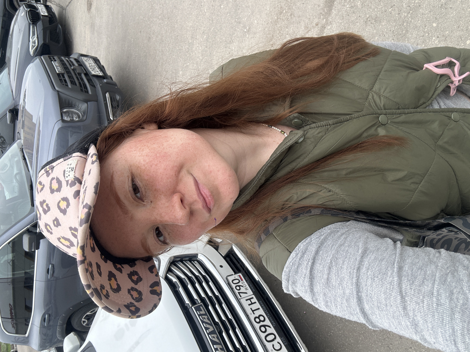A woman is pictured in a selfie-style image next to a white HAVAL vehicle. The car's license plate reads "C098TH 790 RUS," indicating the car is registered in Russia. The photo was taken in Nizhny Novgorod, Russia. The woman is wearing a leopard print hat, a green quilted jacket, and a gray long-sleeved shirt. She has red hair and freckles. Other vehicles are visible in the background, including a gray SUV. The background is a concrete surface, possibly a parking lot or street.